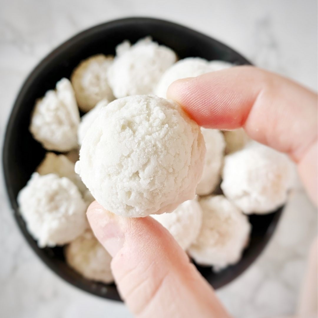 A bowl of coconut fat bombs with a person holding one between their fingers.