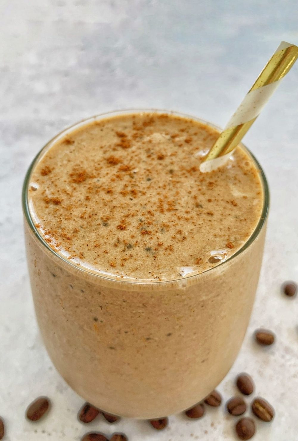 A coffee protein smoothie in a glass with a straw and coffee beans around it.