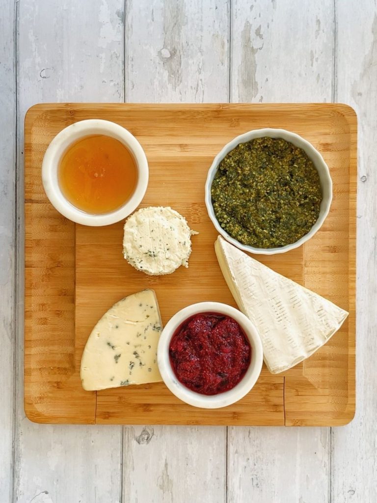 Honey, pesto, and chia seed jam in bowls, as well as 3 types of cheese on a wooded board.