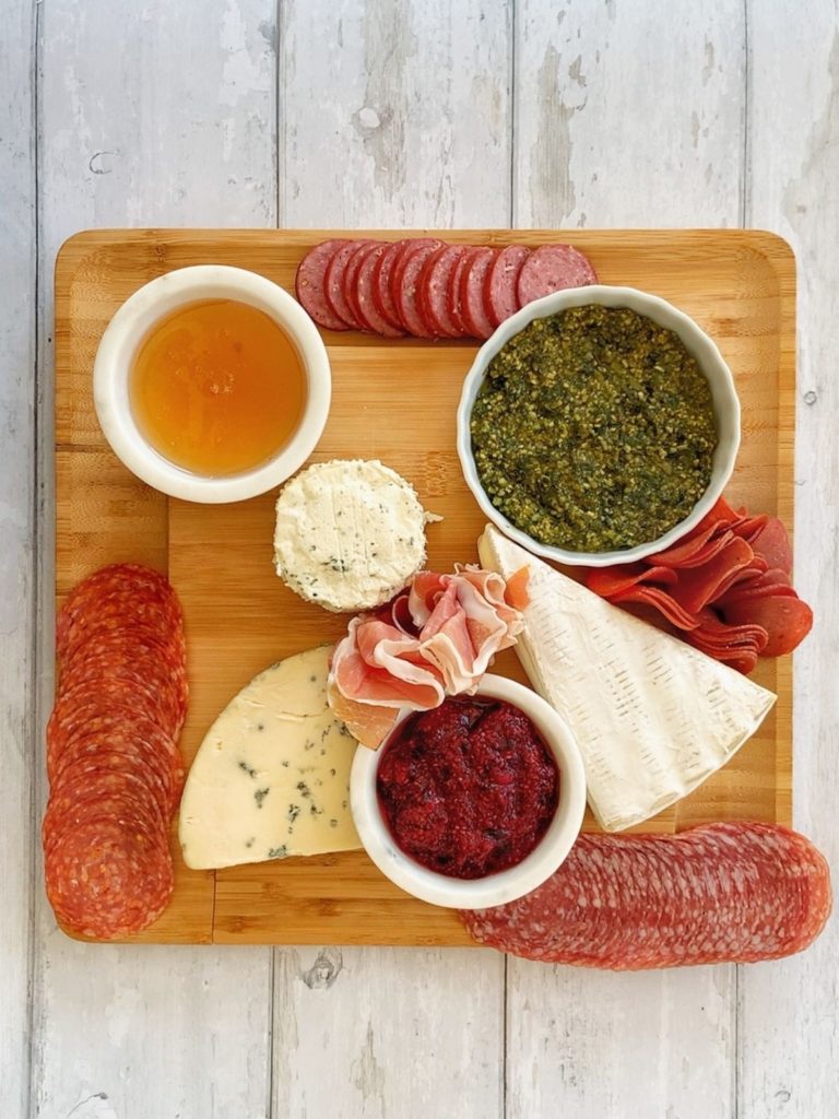 Honey, pesto, chia seed jam, 3 types of cheese, and 5 types of meat on a wooden board.