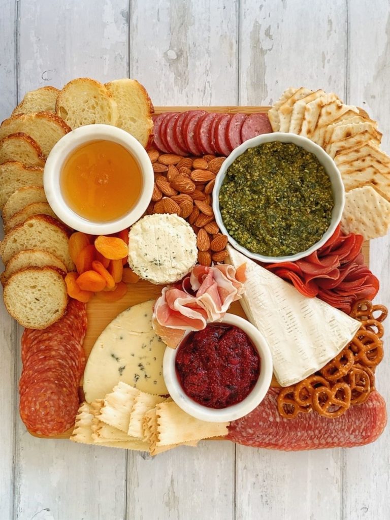 Honey, pesto, chia seed jam, 3 types of cheese, 5 types of meat, crackers, sliced baguette, dried apricots, almonds, and pretzels on a wooden board.
