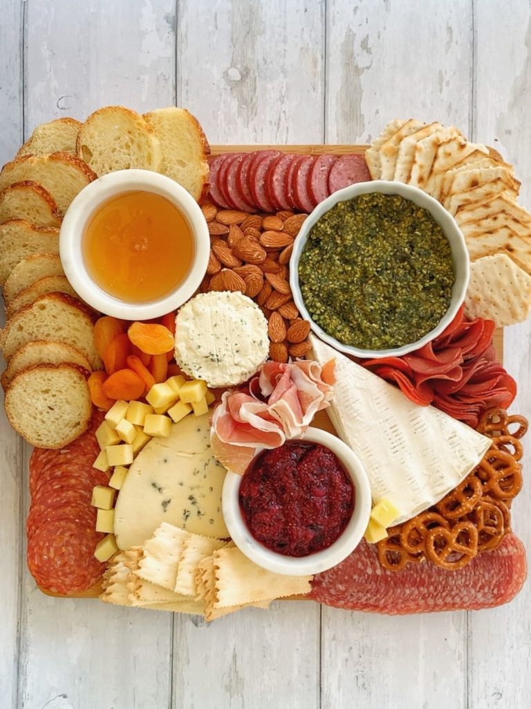 Honey, pesto, chia seed jam, 3 types of cheese, 5 types of meat, crackers, sliced baguette, dried apricots, almonds, pretzels, and cubed cheddar on a wooden board.