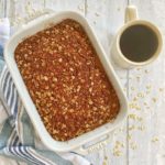 A coffee cake baked in a white pan and topped with cinnamon oat streusel and a cup of coffee.