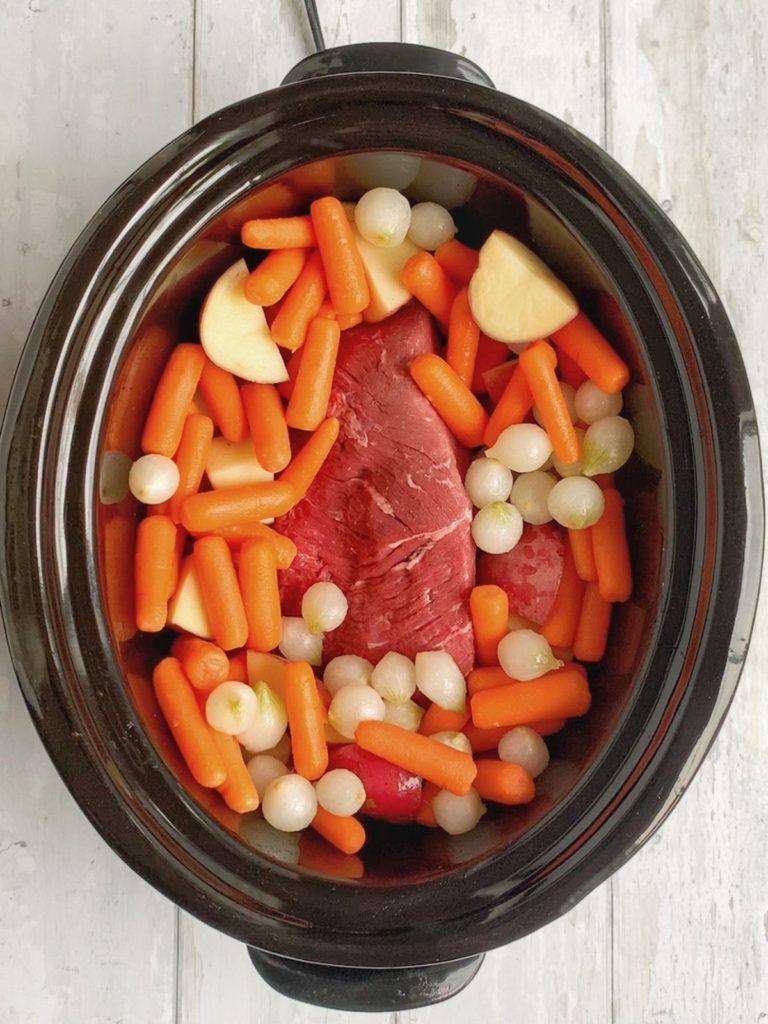A raw rump roast topped with onions, potatoes, and carrots in crock pot.