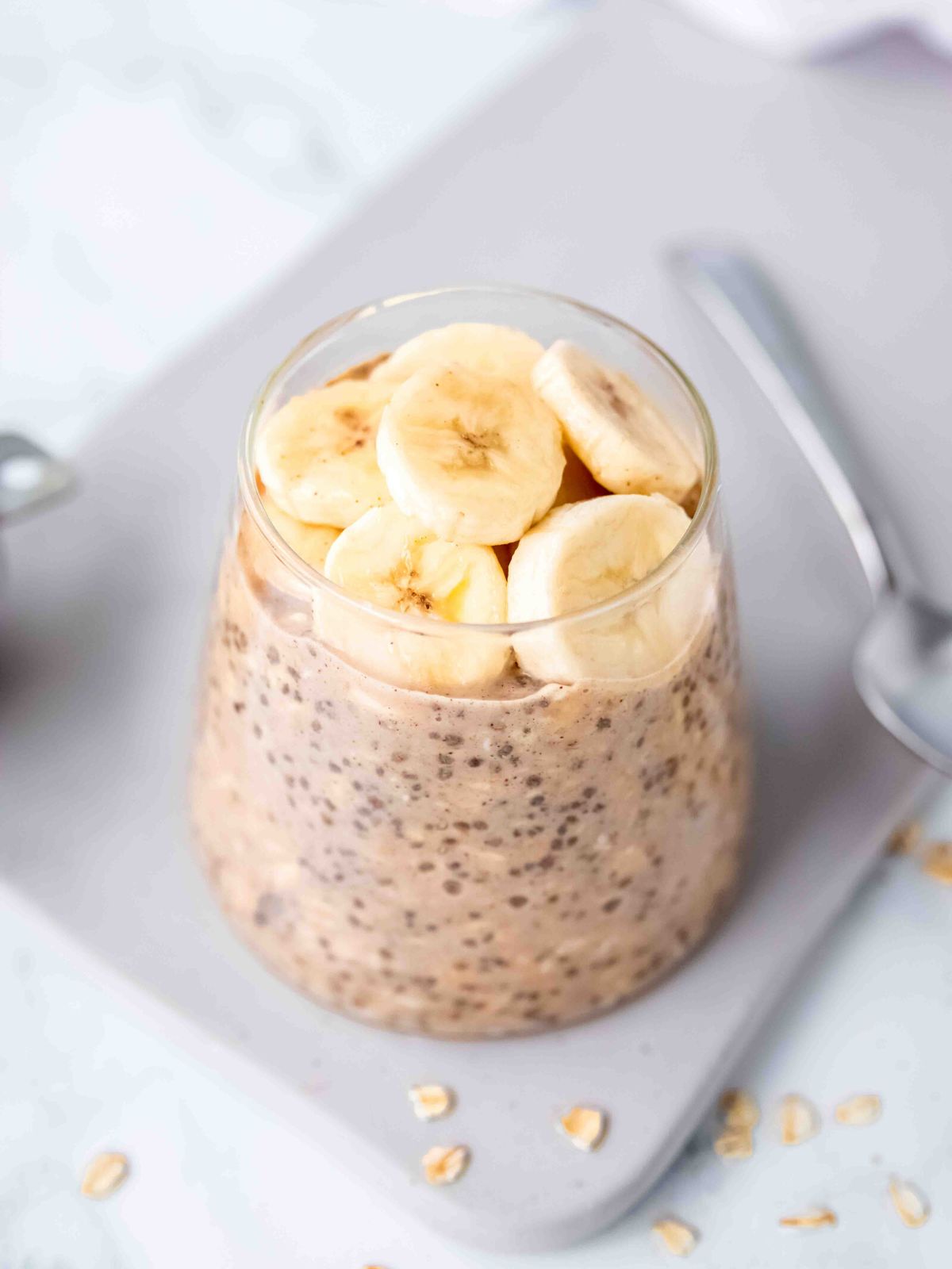 45 degree angle shot of a glass of protein overnight oats topped with banana on a rectangular grey plate with a silver spoon.