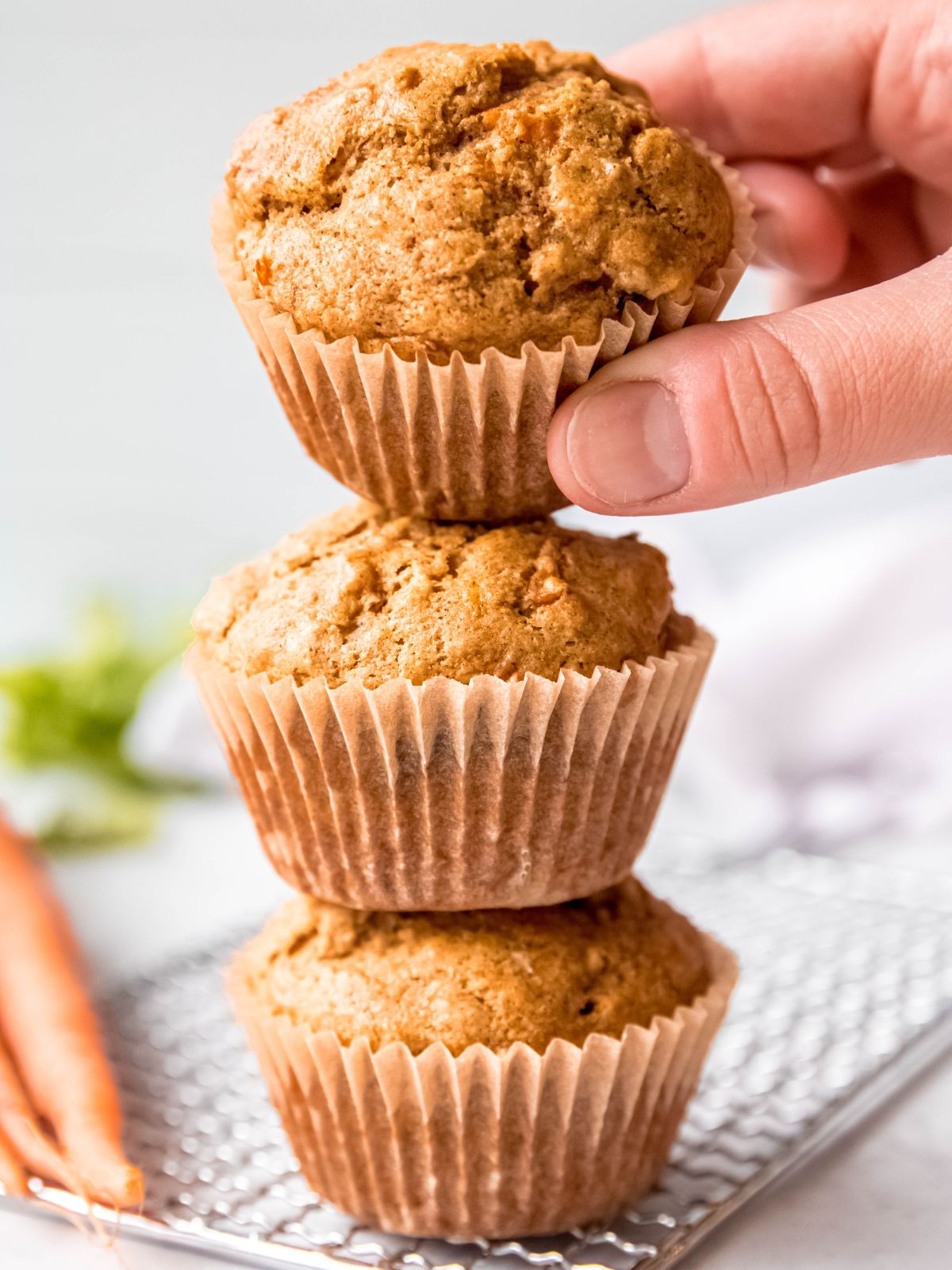 hand pulling the top muffin from a stack of 3 carrot banana muffins on a cooling rack.