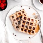 square hero image of 2 whole wheat chocolate chip waffles dusted with powdered sugar and drizzled with maple syrup on a white plate.