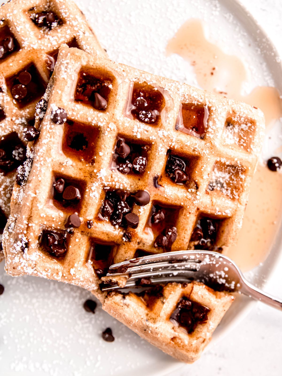 hero shot of a fork taking a bite out of a chocolate chip waffle drizzled with maple syrup and sprinkled with powdered sugar.