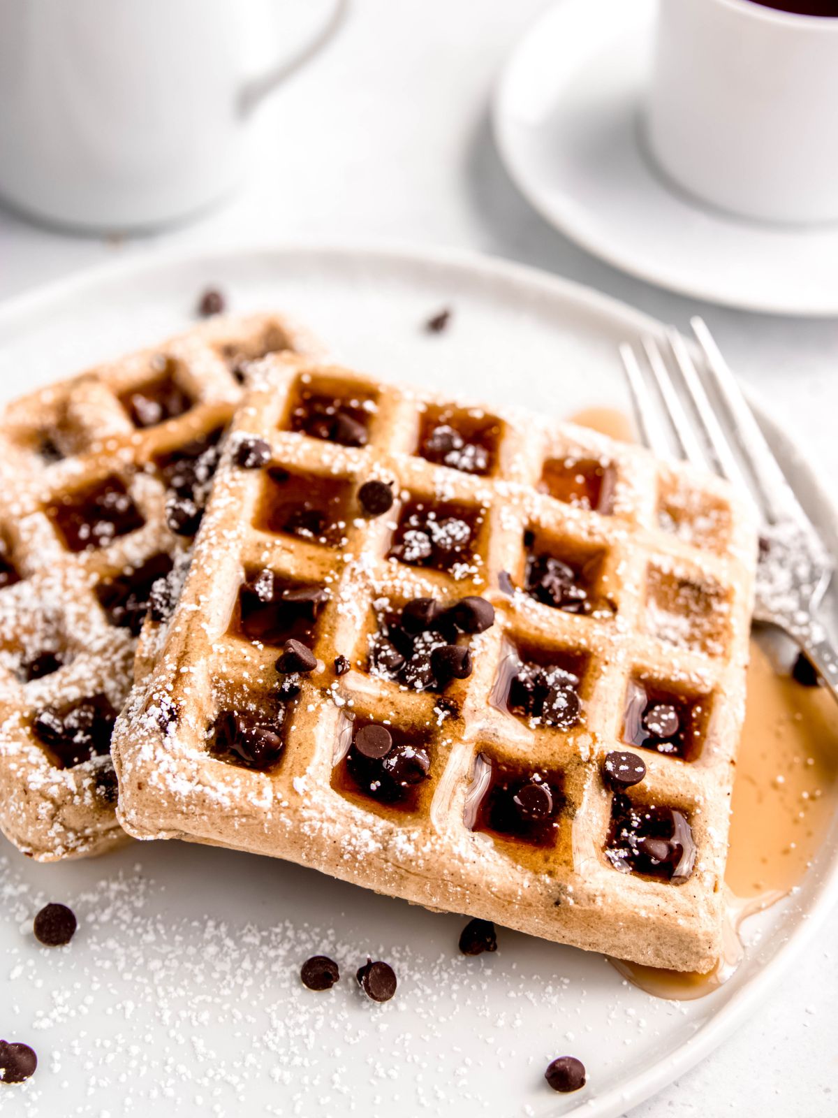 45 degree angle shot of two chocolate chip waffles topped with extra chocolate chips, maple syrup and powdered sugar on a white plate with a silver fork.