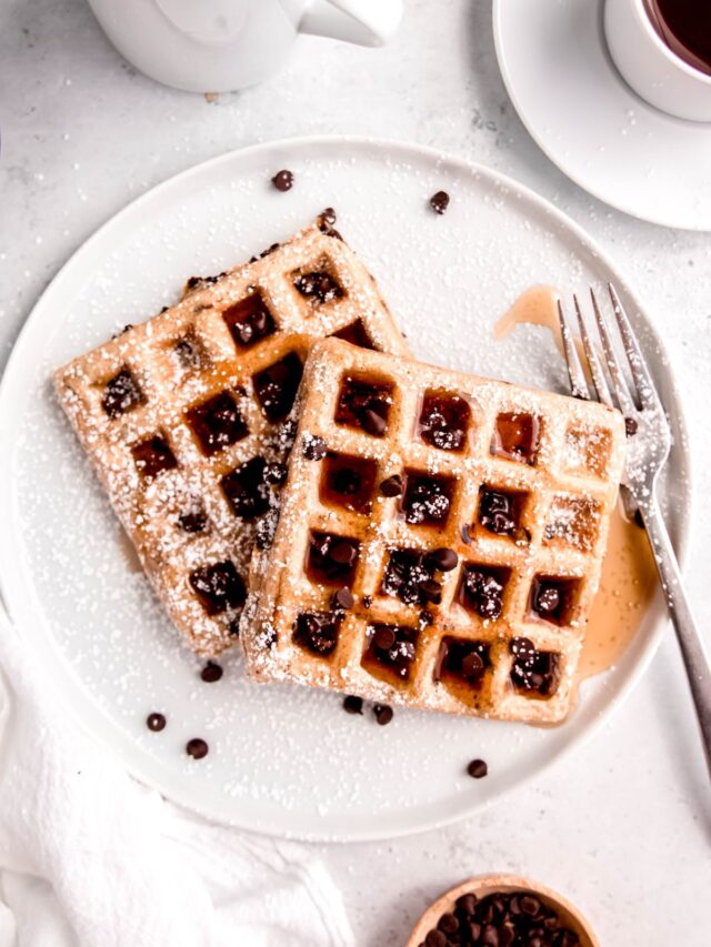 flat lay shot of two chocolate chip waffles topped with extra chocolate chips, maple syrup and powdered sugar on a white plate with a silver fork.