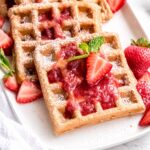 square hero image of homemade strawberry waffles on a white plate, topped with powdered sugar, strawberry syrup, fresh strawberries, and mint.