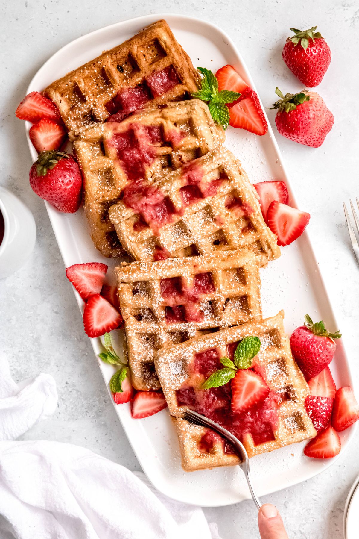 5 homemade strawberry waffles on a rectangular white serving platter topped with strawberry waffle syrup, fresh strawberries, and powdered sugar.