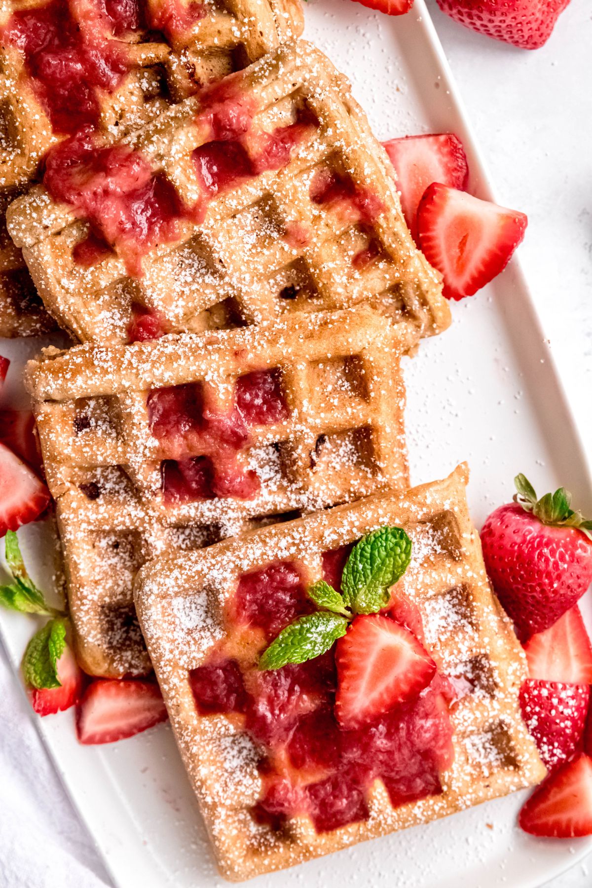 overhead shot of a serving platter of strawberry waffles topped with strawberry waffle syrup, fresh strawberry slices, powdered sugar, and fresh mint.