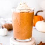 square hero image of a pumpkin protein shake in a clear glass topped with whipped cream and pumpkin pie spices.