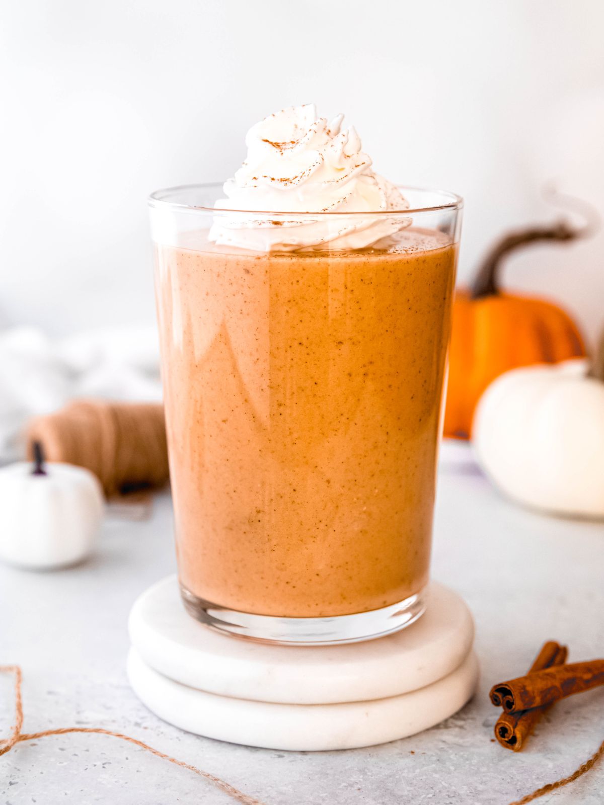 side on hero image of a clear glass filled with pumpkin protein shake, topped with whipped cream, on a table with mini pumpkins and cinnamon sticks.