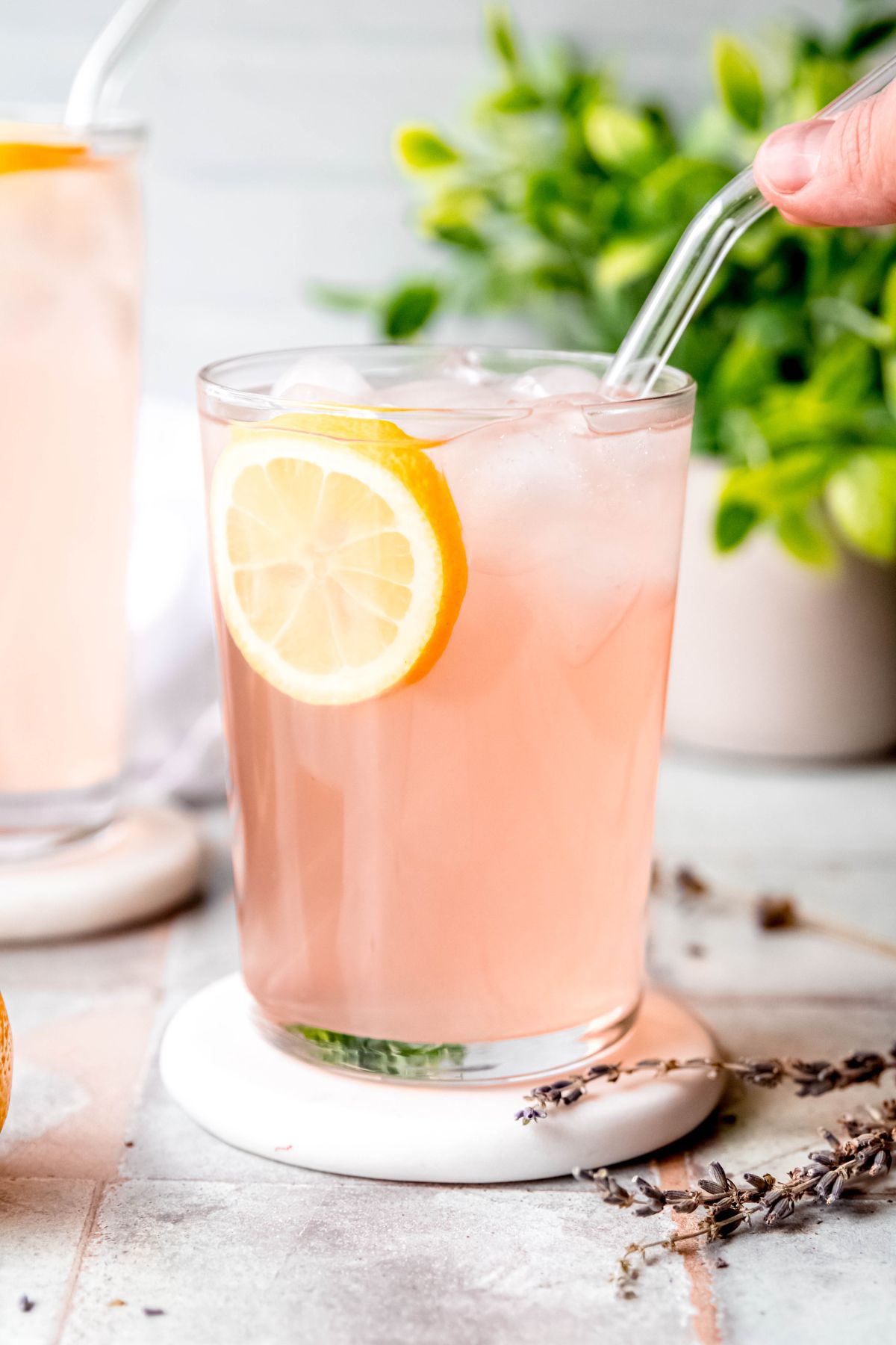 side on view of a glass of homemade lavender lemonade with a round of lemon on a white coaster with a sprig of lavender in the foreground.