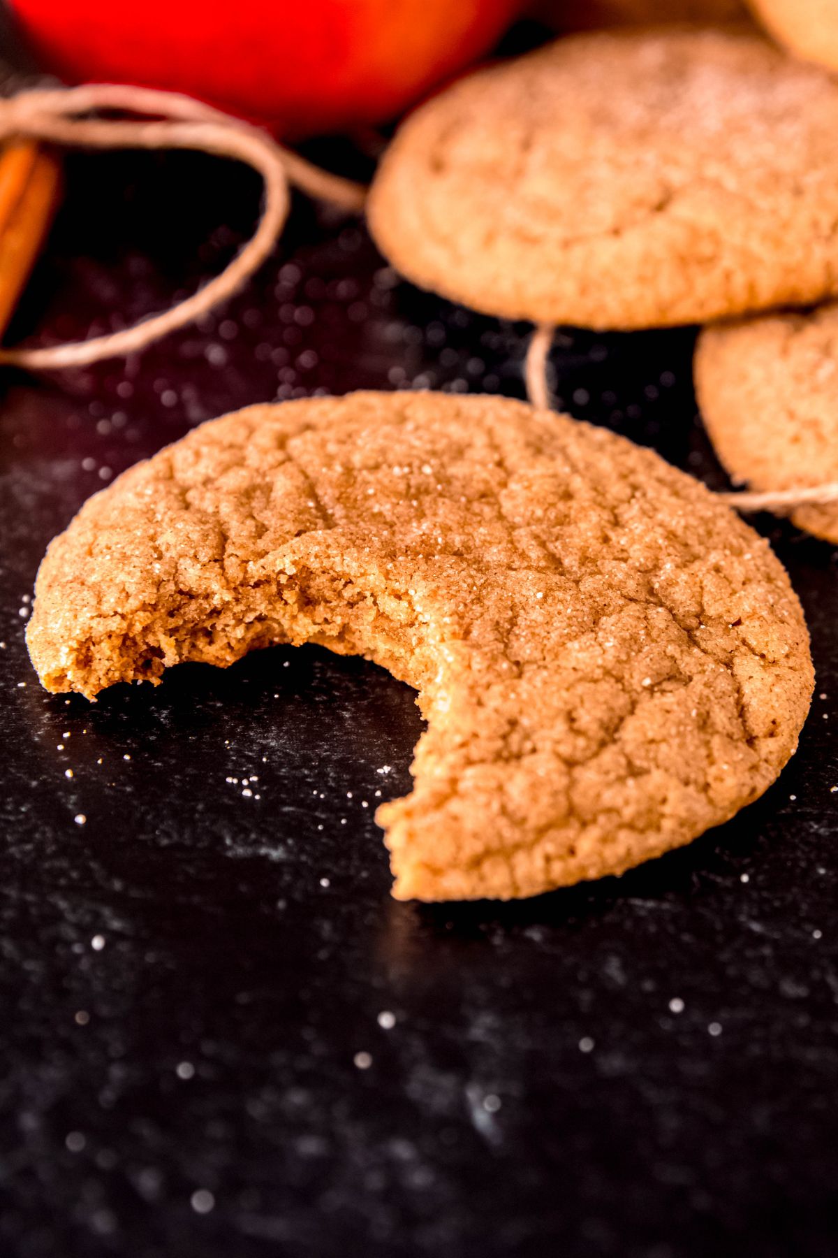 45 degree angle shot of a freshly baked apple cider cookie with a bite taken out of it on a black table with cinnamon sugar scattered around and more cookies, twine, and a whole apple blurred out in the background.