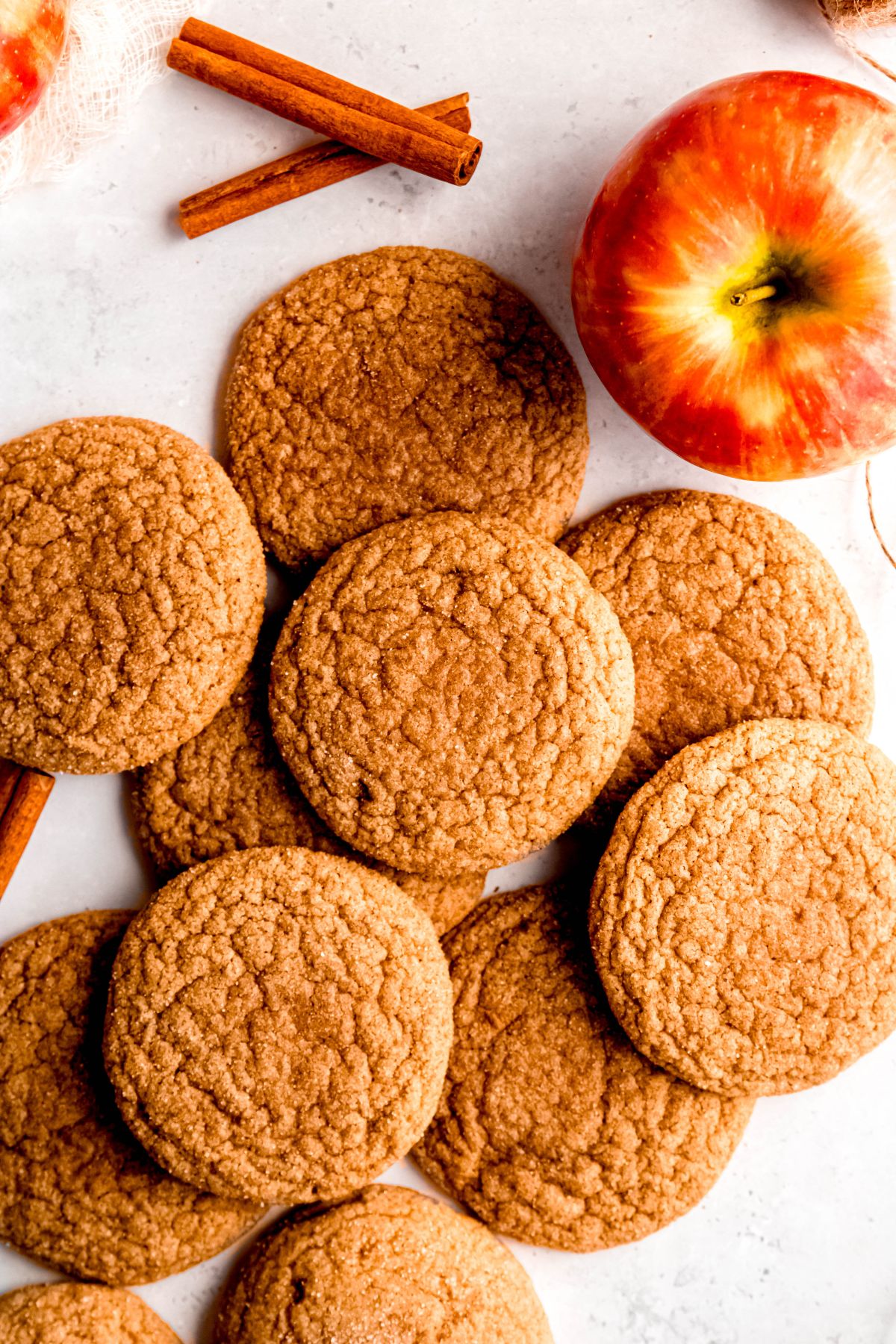flat lay shot of a messy pile of freshly baked spiced apple cider cookies on a white table with whole cinnamon sticks and a whole apple.