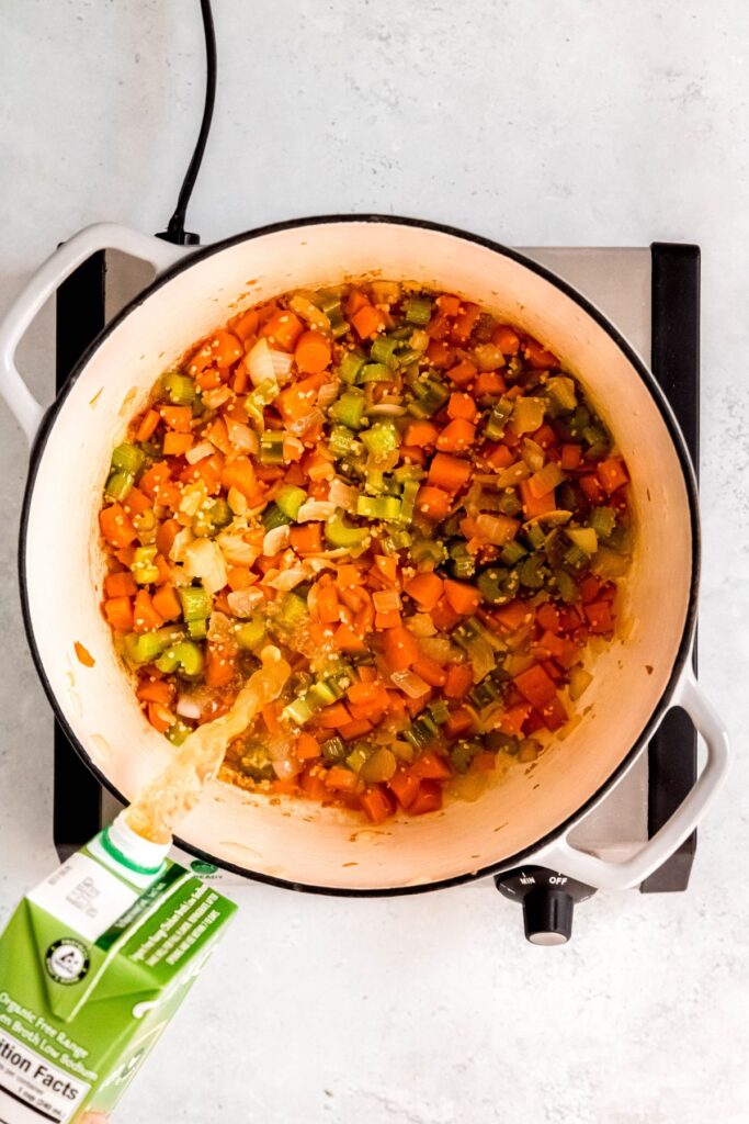 action shot of chicken broth being poured into a white dutch oven with sautéed mirepoix and garlic mixture.