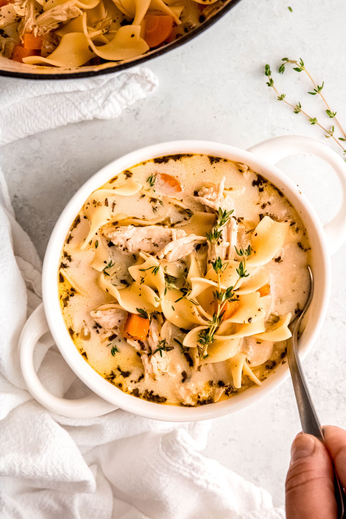 hand grabbing a silver soup spoon handle that is dunked into a small white soup crock with two handles filled with creamy turkey noodle soup on a white table with a white Dutch oven with the rest of the soup in the top of the frame with a few sprigs of fresh thyme.