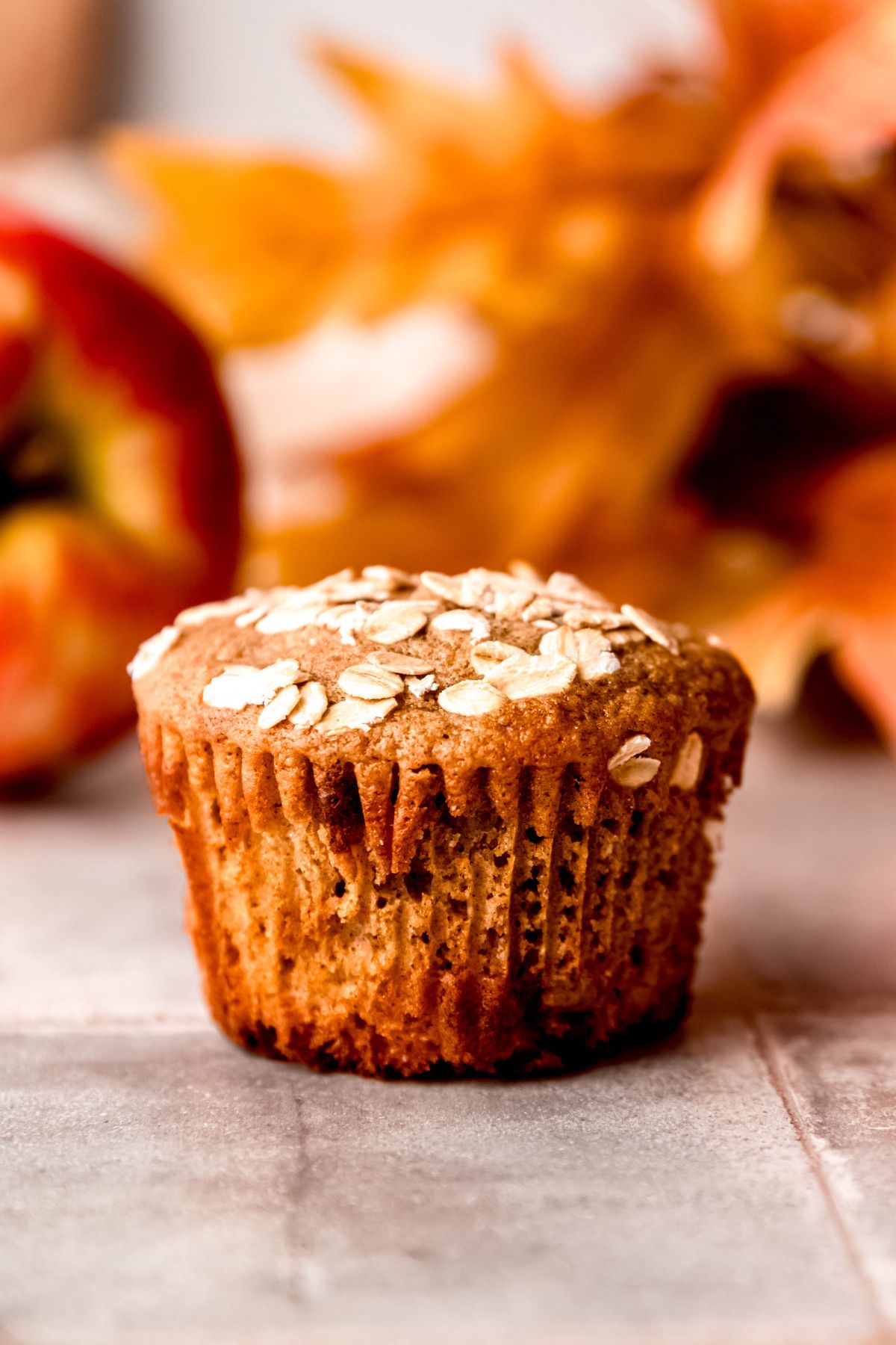 A side shot of an apple cinnamon protein muffin on the counter with apples and leaves in the background.