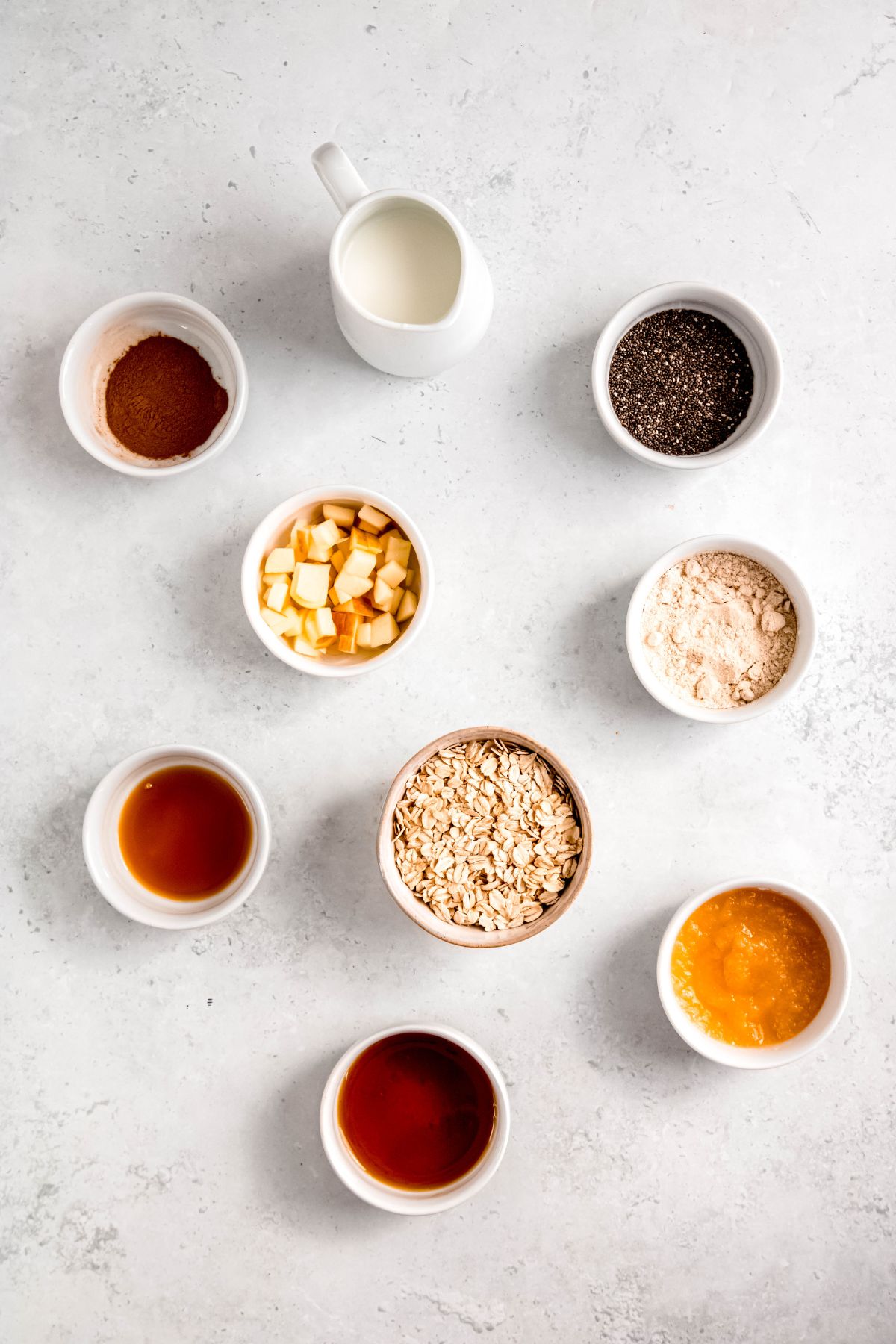 ingredients for making high-protein apple cinnamon overnight oats with protein powder all measured out into white bowls on a white table.