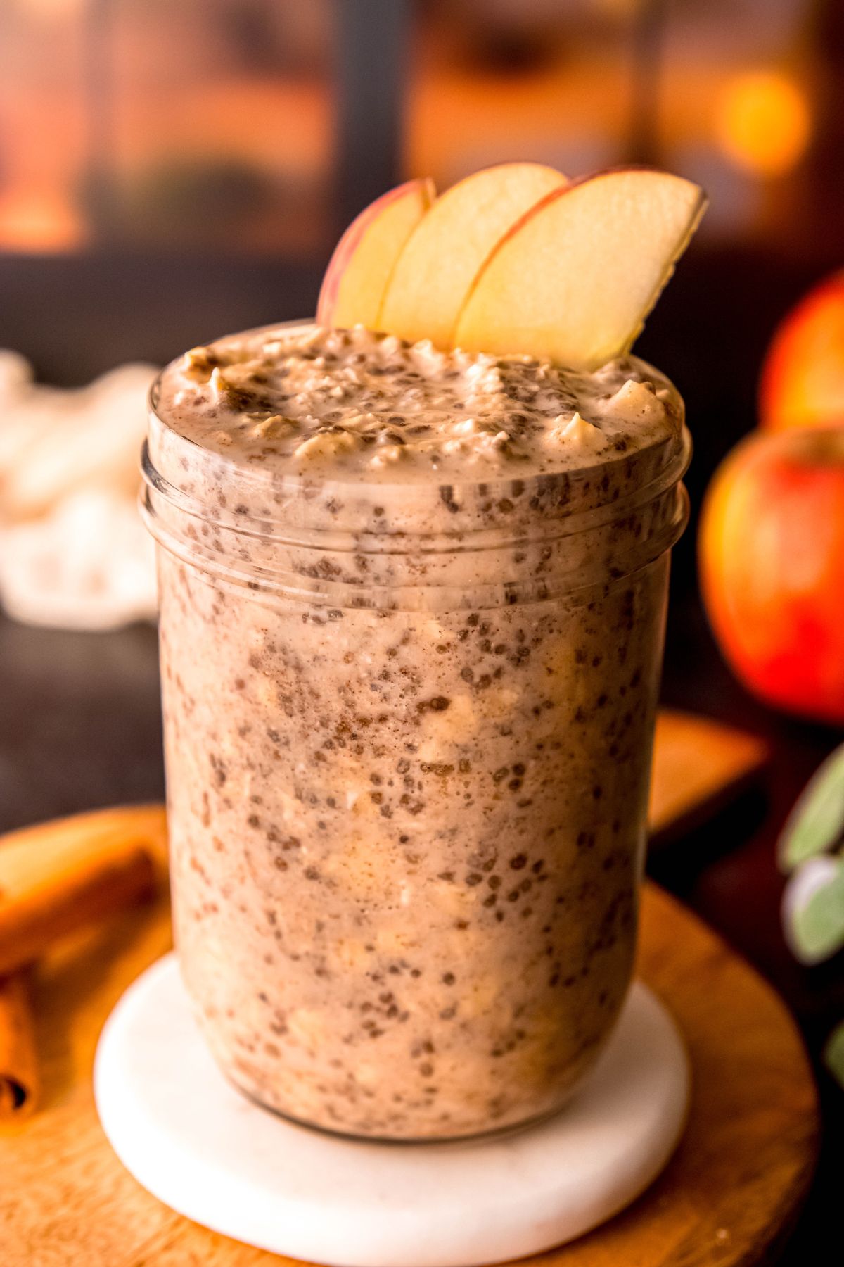 side on view of a mason jar of protein overnight oats with apples, cinnamon and chia seeds, garnished with 3 slices of fresh apple and an extra sprinkling of cinnamon in front of a cozy window lit by fairy lights.