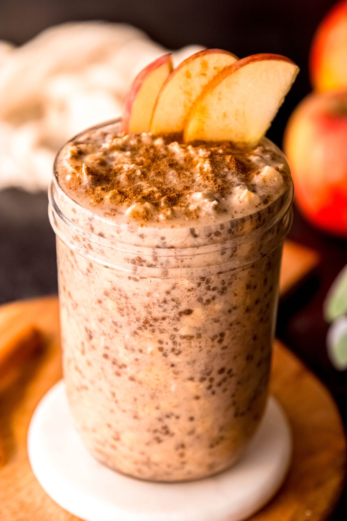 jar of apple cinnamon protein overnight oats garnished with 3 thin slices of apples and a sprinkle of cinnmaon on a wooden tray with whole cinnamon sticks, and an apple, green houseplant, and white cloth blurred out behind.