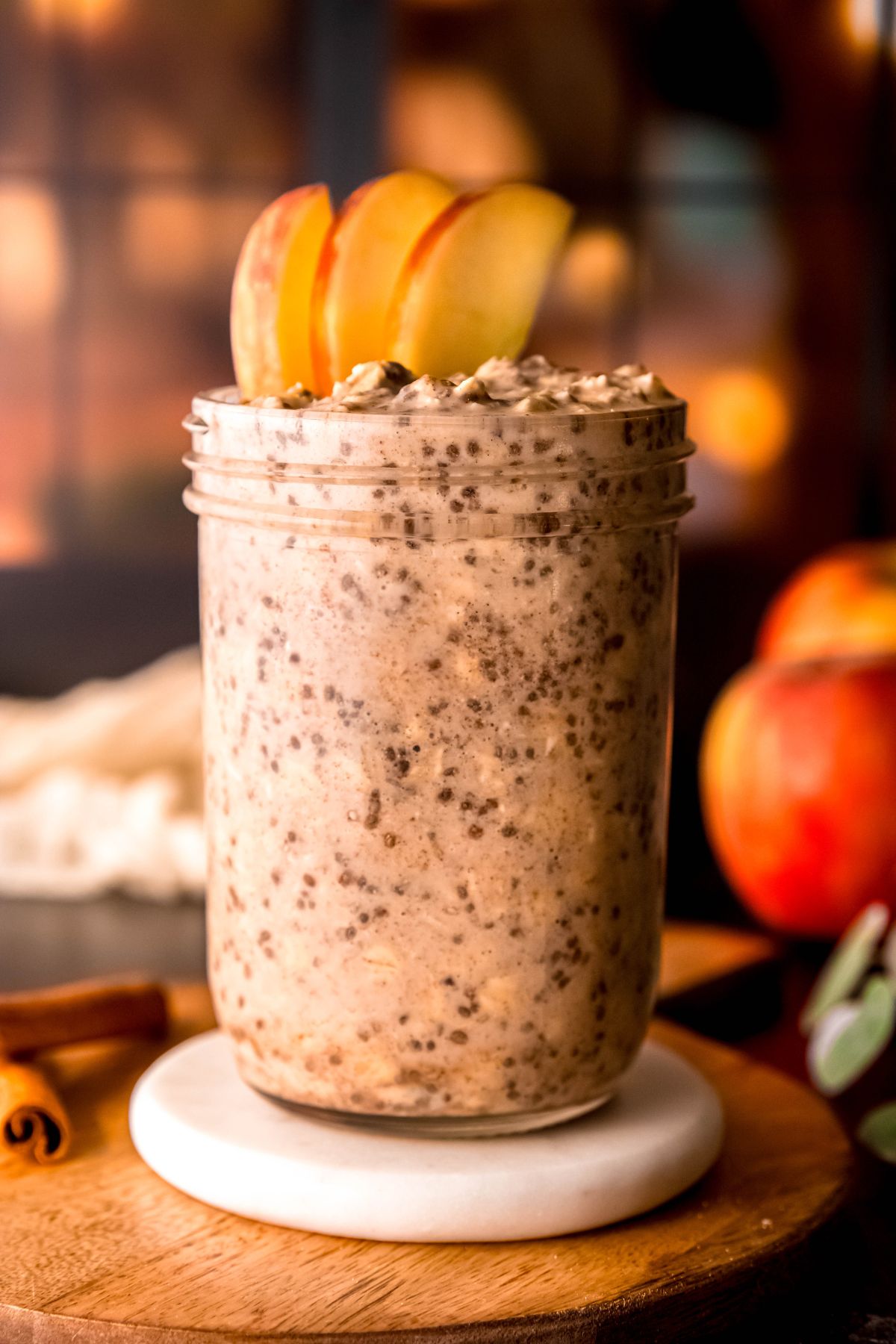 hero shot of a mason jar of overnight cinnamon apple oats with protein powder topped with apple slices and extra cinnamon on a white coaster on a wooden table with two cinnamon sticks stacked to the left and apples blurred out in the background in front of a window with fairy lights shining through.
