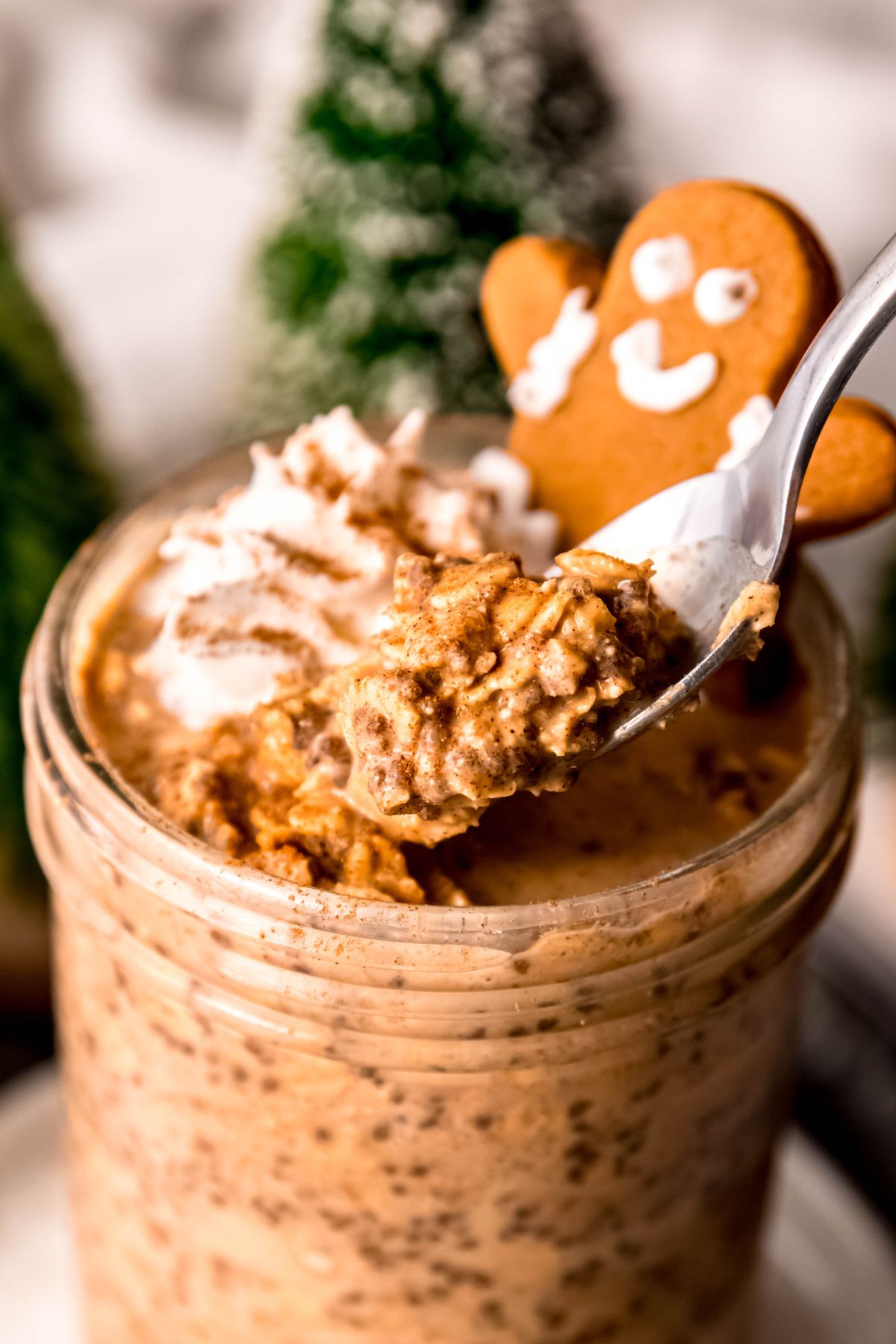 closeup on a silver spoonful of gingerbread flavored protein overnight oats showing their chewy texture with the rest of the jar and the gingerbread man topper blurred in the background.