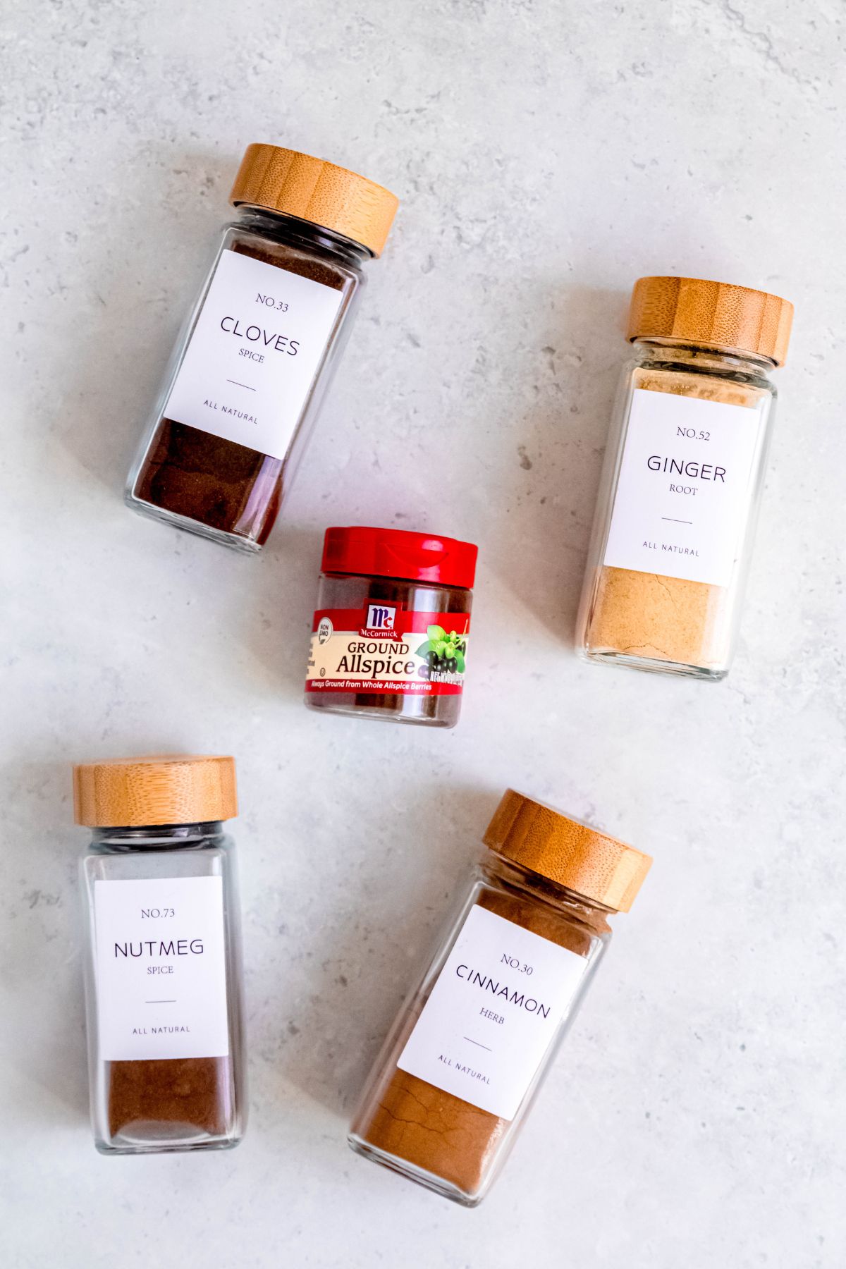 spice bottles filled with cloves, ginger, allspice, nutmeg, and cinnamon laid out on a white table for making DIY pumpkin spice.