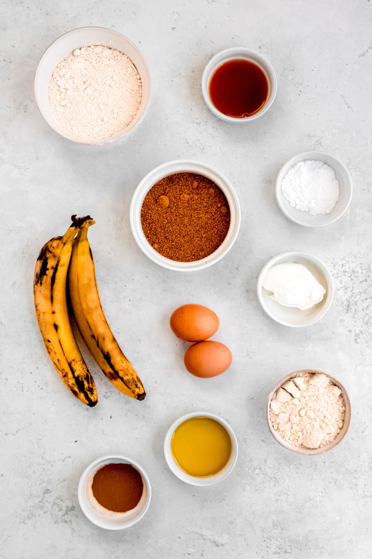 ingredients to make high protein banana bread muffins measured out into white bowls on a grey-white table.