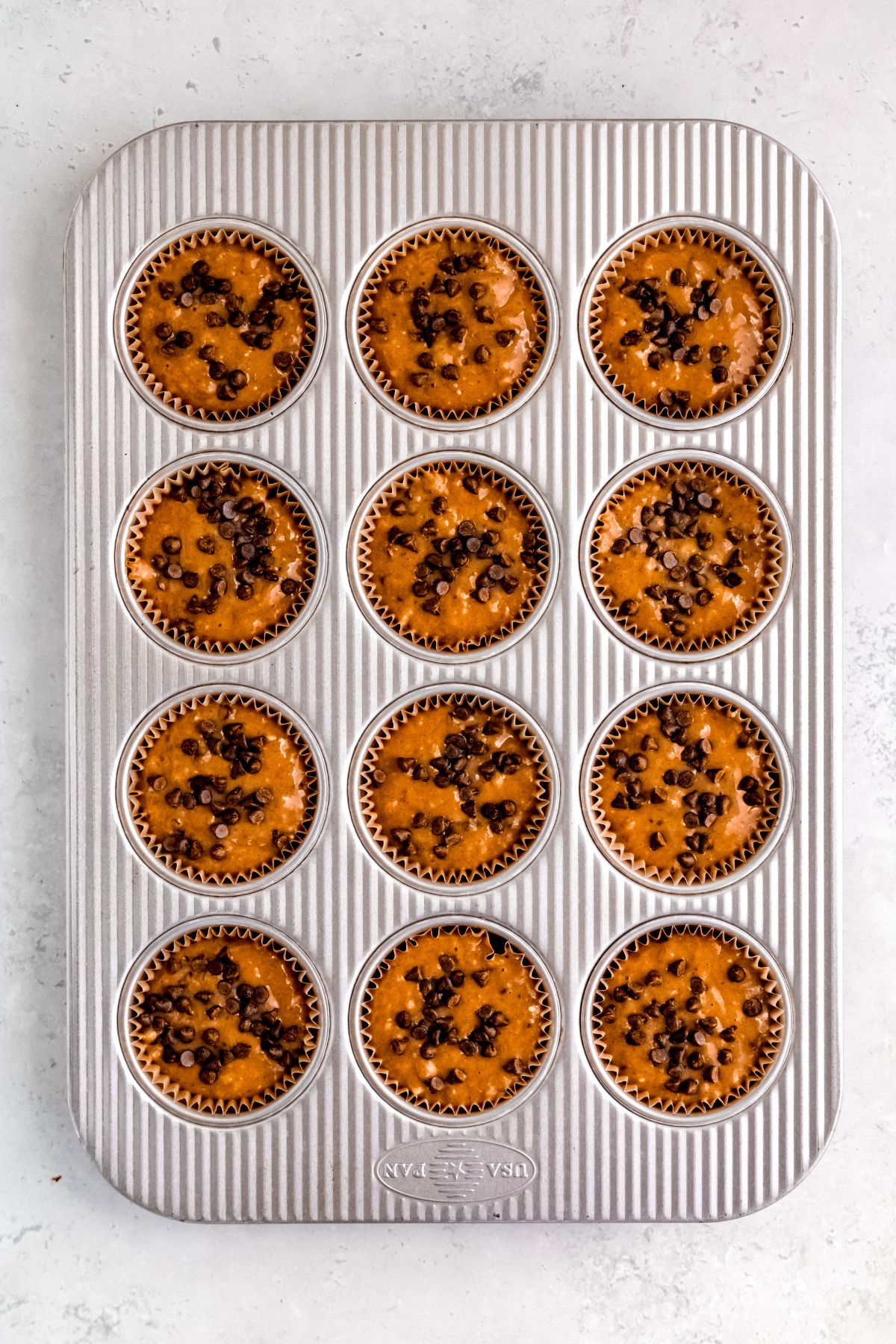 chocolate chip banana protein muffin batter divvied up between 12 muffin wells and sprinkled with extra mini chocolate chips.