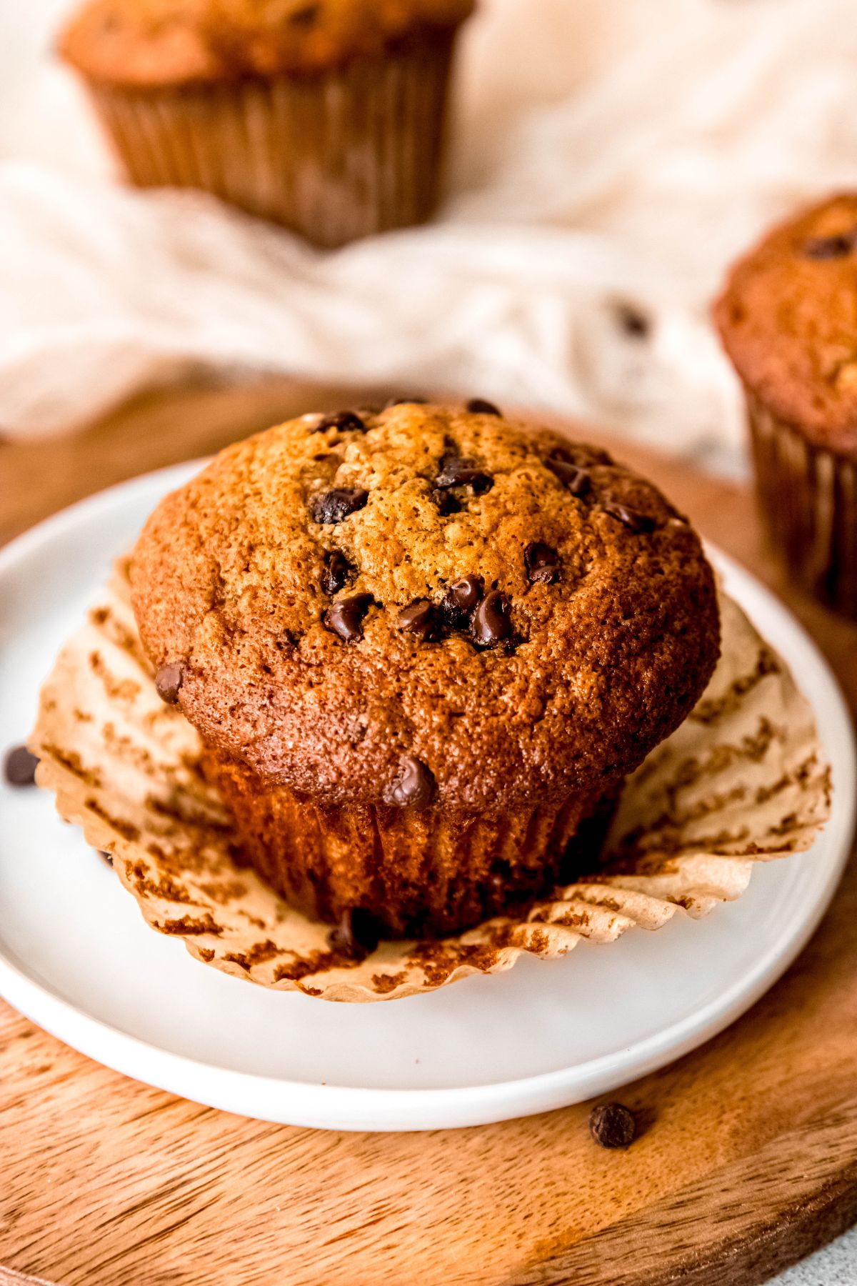 45 degree angle shot of a banana chocolate chip protein muffin with the muffin wrapper peeled away and fanned out on top of a small white round plate on a wooden table with chocolate chips scattered around.