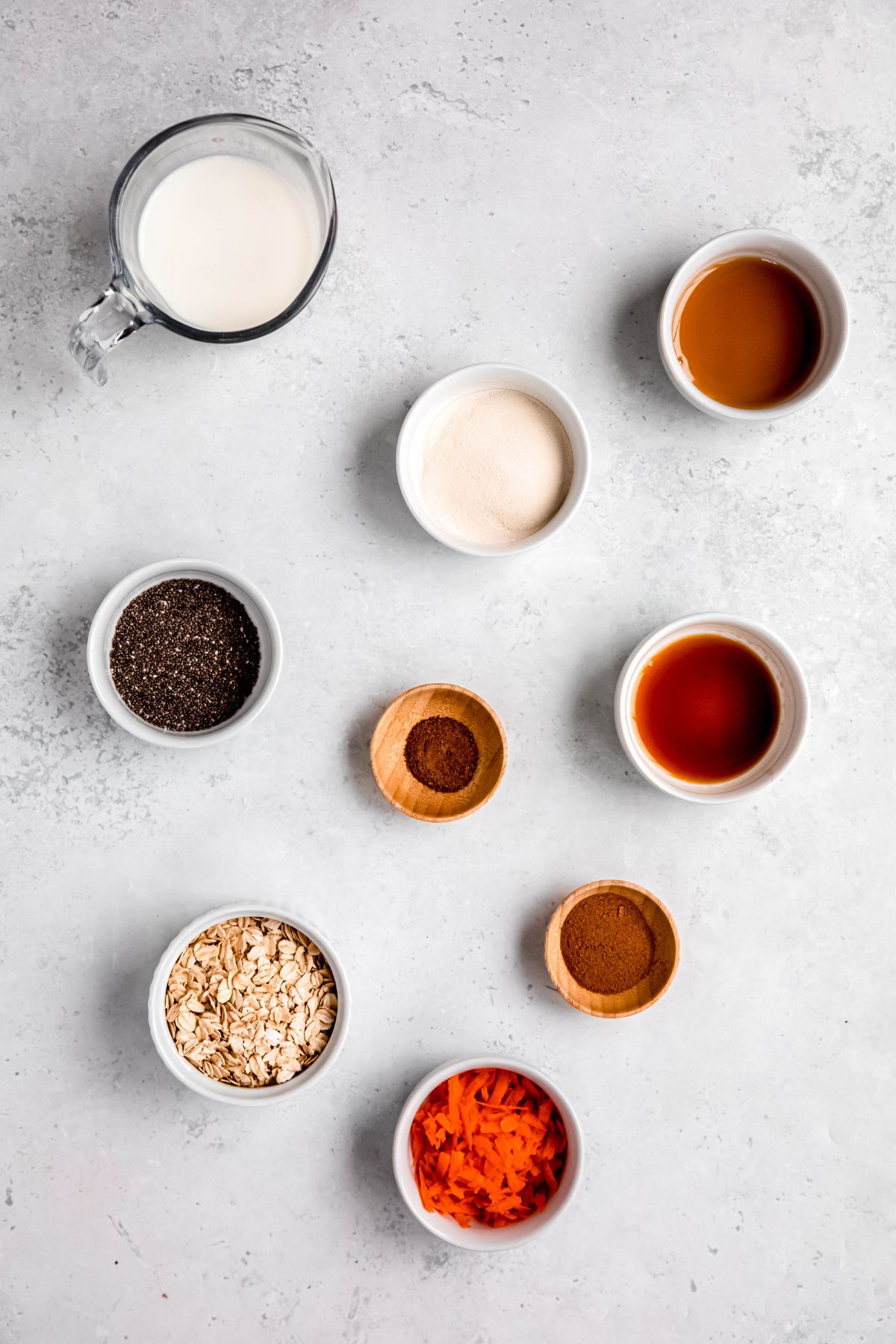 flat lay shot of the ingredients needed to make high protein carrot cake overnight oats measured out into small bowls on a white table.