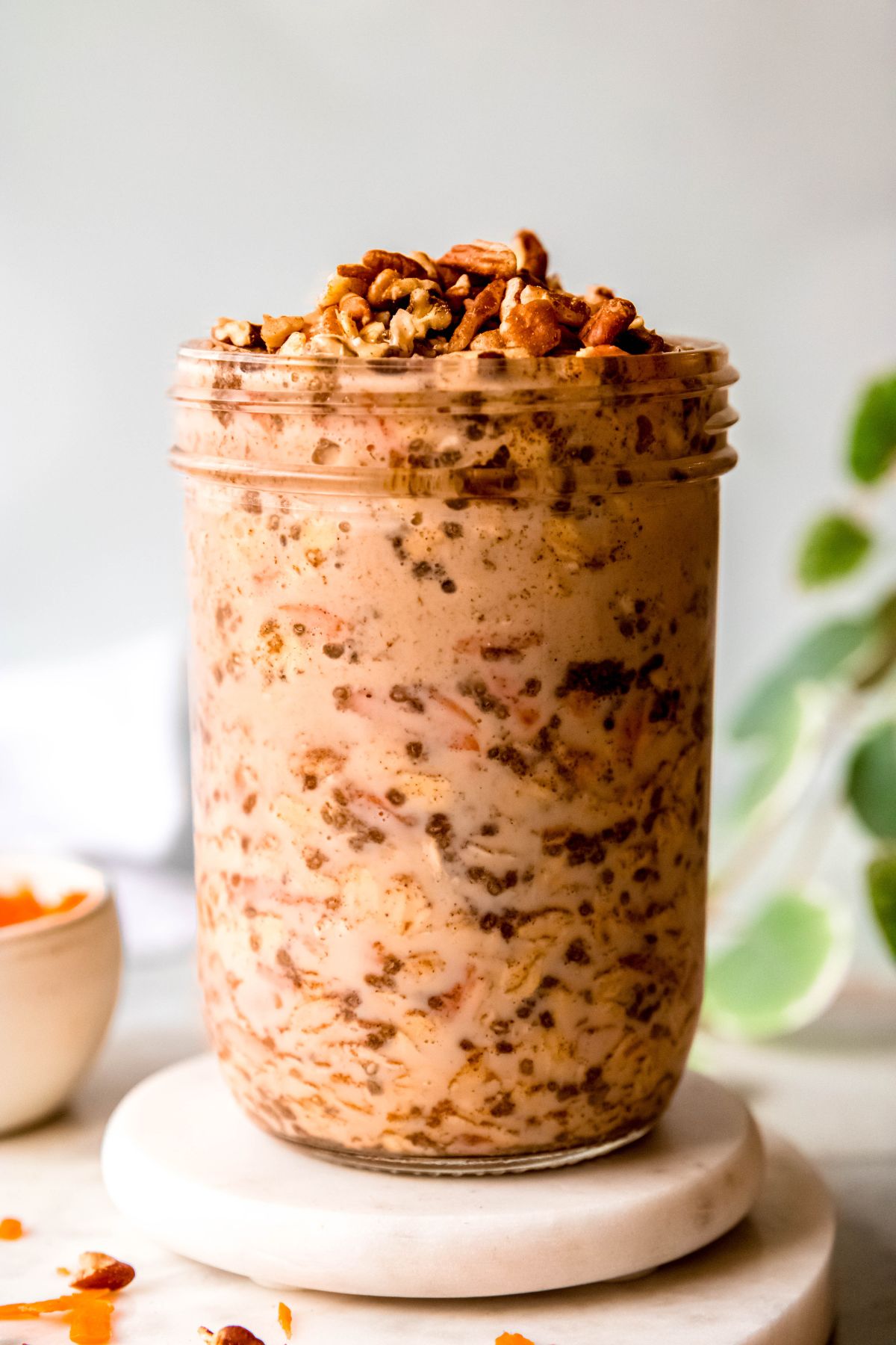 closeup side-on view of a clear glass mason jar of carrot cake overnight oats garnished with nuts and raisins on a white table with a green houseplant blurred out to the right and a white bowl with shredded carrots blurred out on the left.