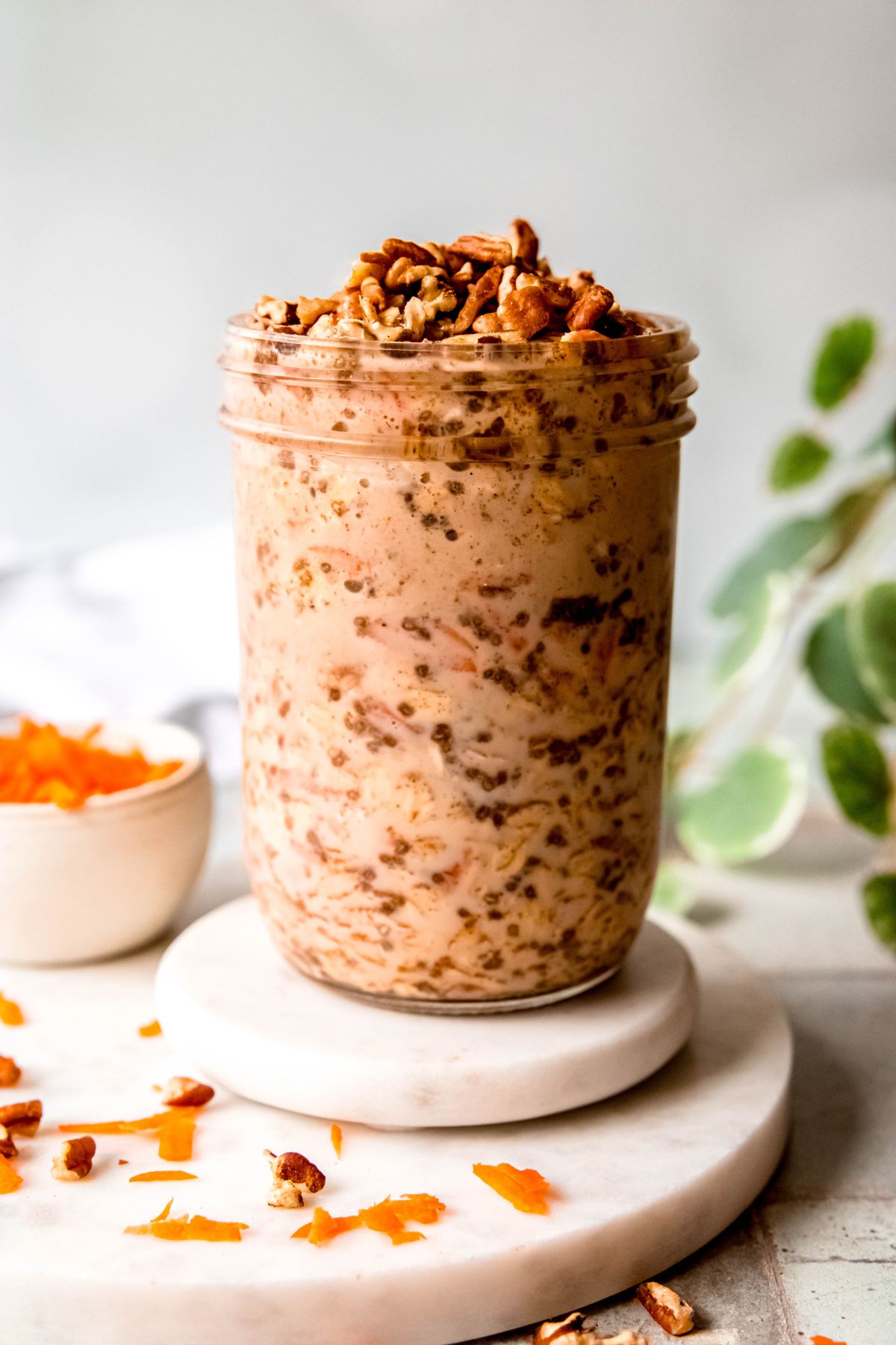 side on view of a mason jar of protein carrot cake overnight oats on a white coaster atop a round white marble serving tray with carrot shreds and chopped pecans sprinkled around.