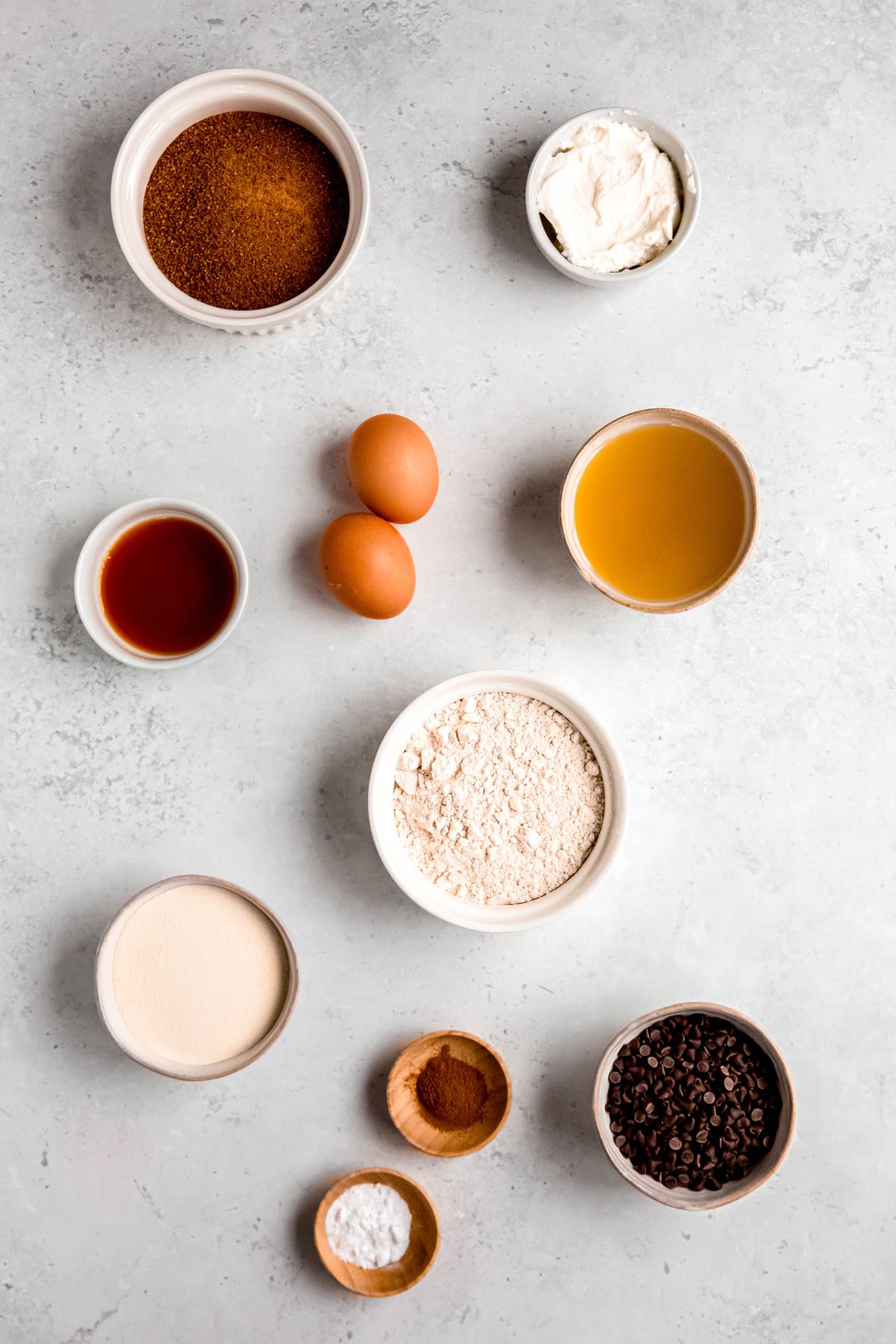 ingredients needed to make healthy chocolate chip muffins with protein powder and whole wheat measured out into small white bowls on a white table.