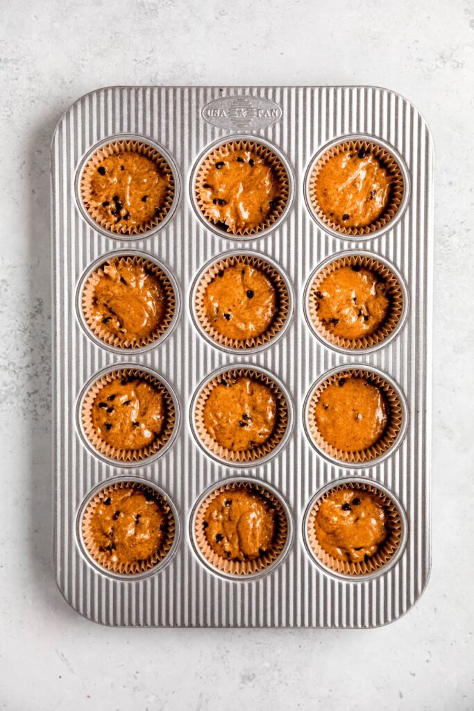 chocolate chip protein powder muffin batter dished out into a silver 12-well muffin tin lined with paper liners.