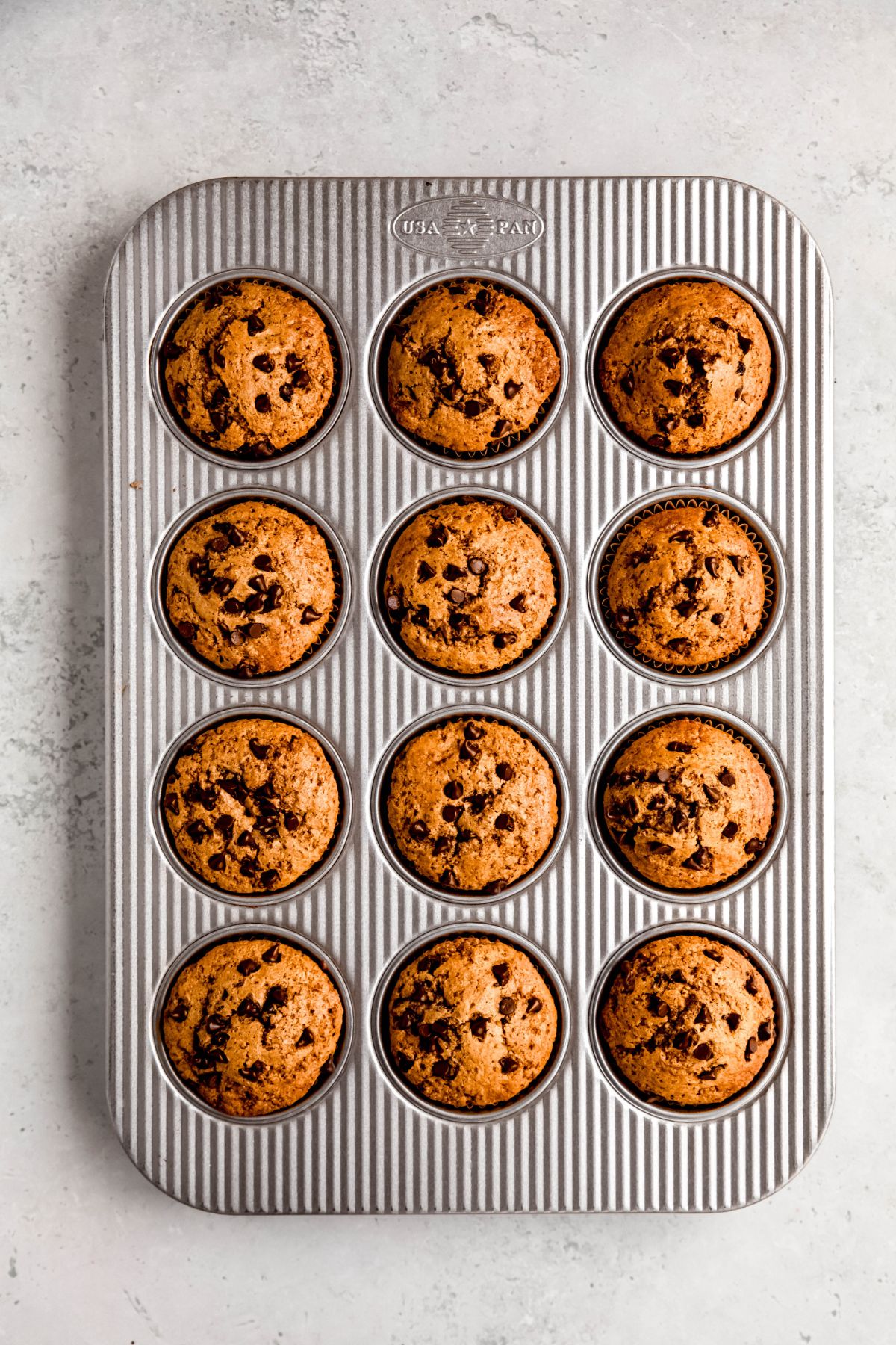 flat lay shot of baked protein chocolate chip muffins in a silver baking tin on top of a pale grey table.