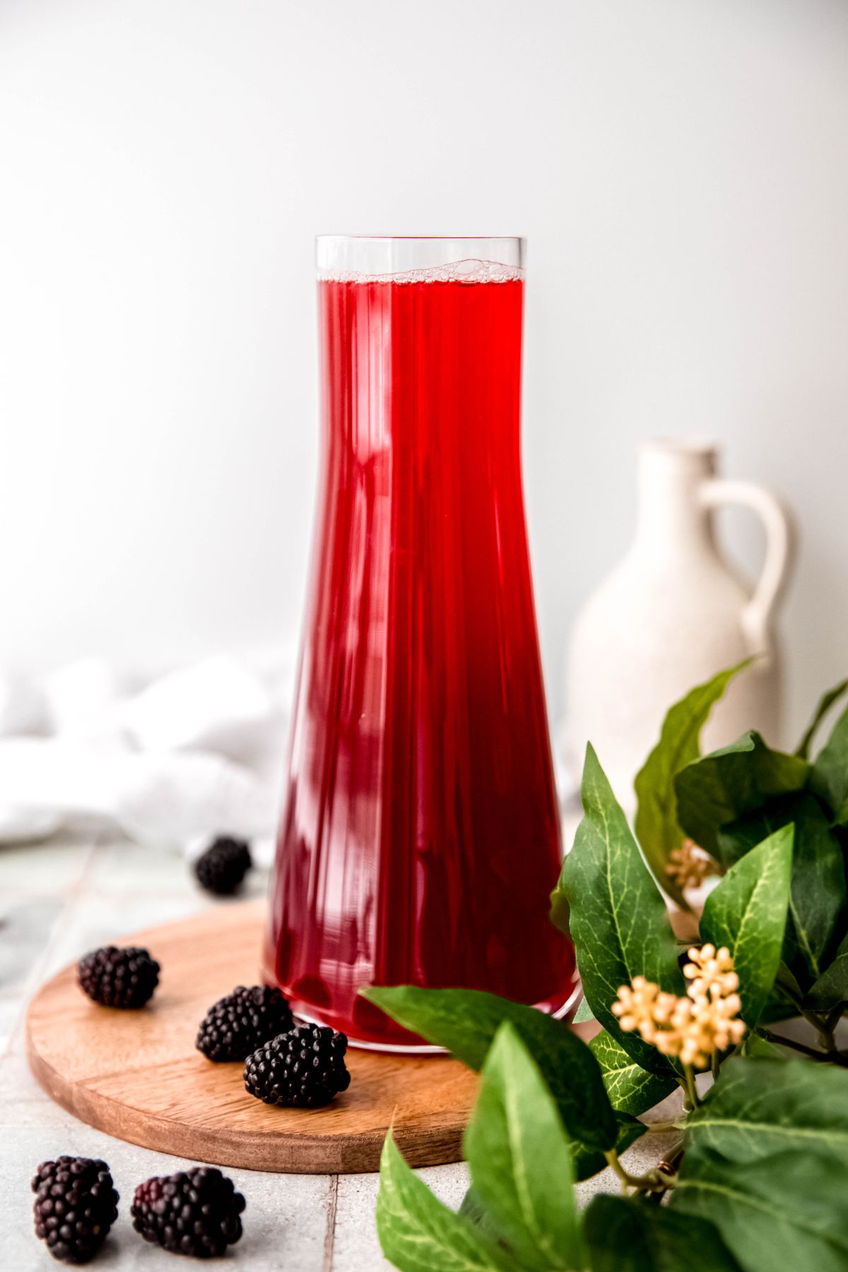 clear glass handle-less pitcher filled with homemade blackberry lemonade on a wooden serving tray on top of a grey tile table with blackberries scattered around and a green houseplant.