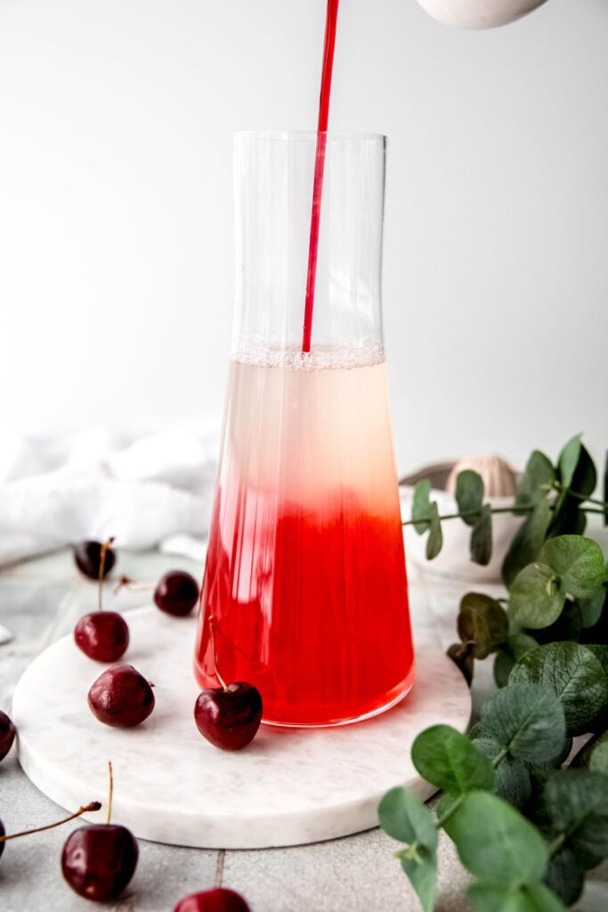 cherry syrup being poured into the lemon-water mixture; the syrup is making the bottom of the pitcher red, while the murky yellow lemon water is separated on top before stirring.