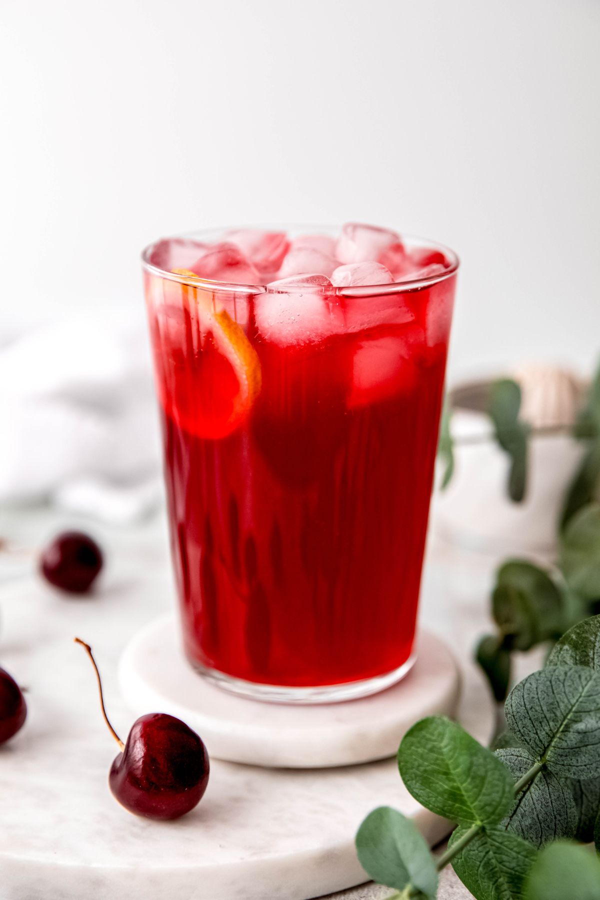 easy homemade cherry lemonade in a clear glass on a white coaster on a white table surrounded by sprigs of a green houseplant and fresh cherries.