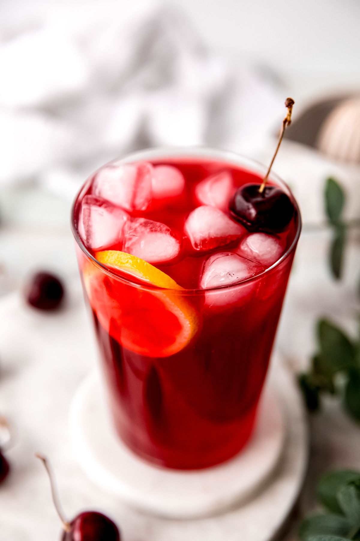45 degree angle shot looking down into a glass of cherry lemonade on a white table with a green houseplant and fresh black cherries blurred out beneath.