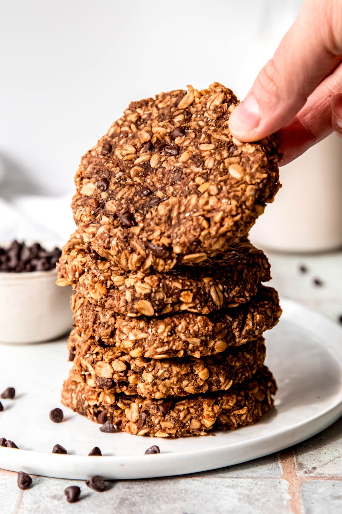 hand grabbing the top chocolate protein oatmeal cookie from a stack of 5 cookies on a white plate with a white bowl of mini chocolate chips and a white milk jug blurred out in the background.