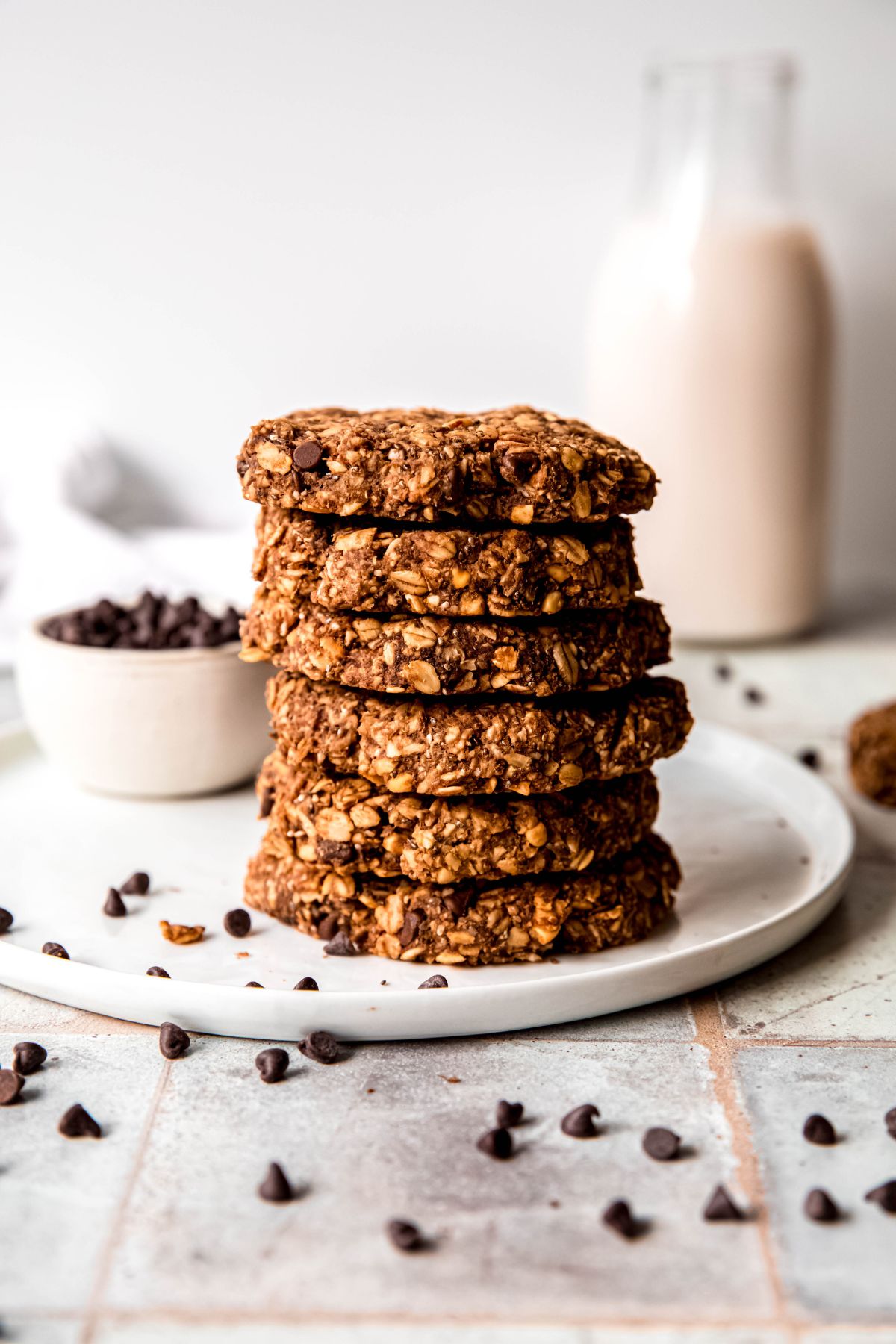 stack of 6 chocolate oatmeal protein cookies on a round white plate with a small white bowl of mini chocolate chips on it in the background and a smattering of mini chocolate chips scattered all around.