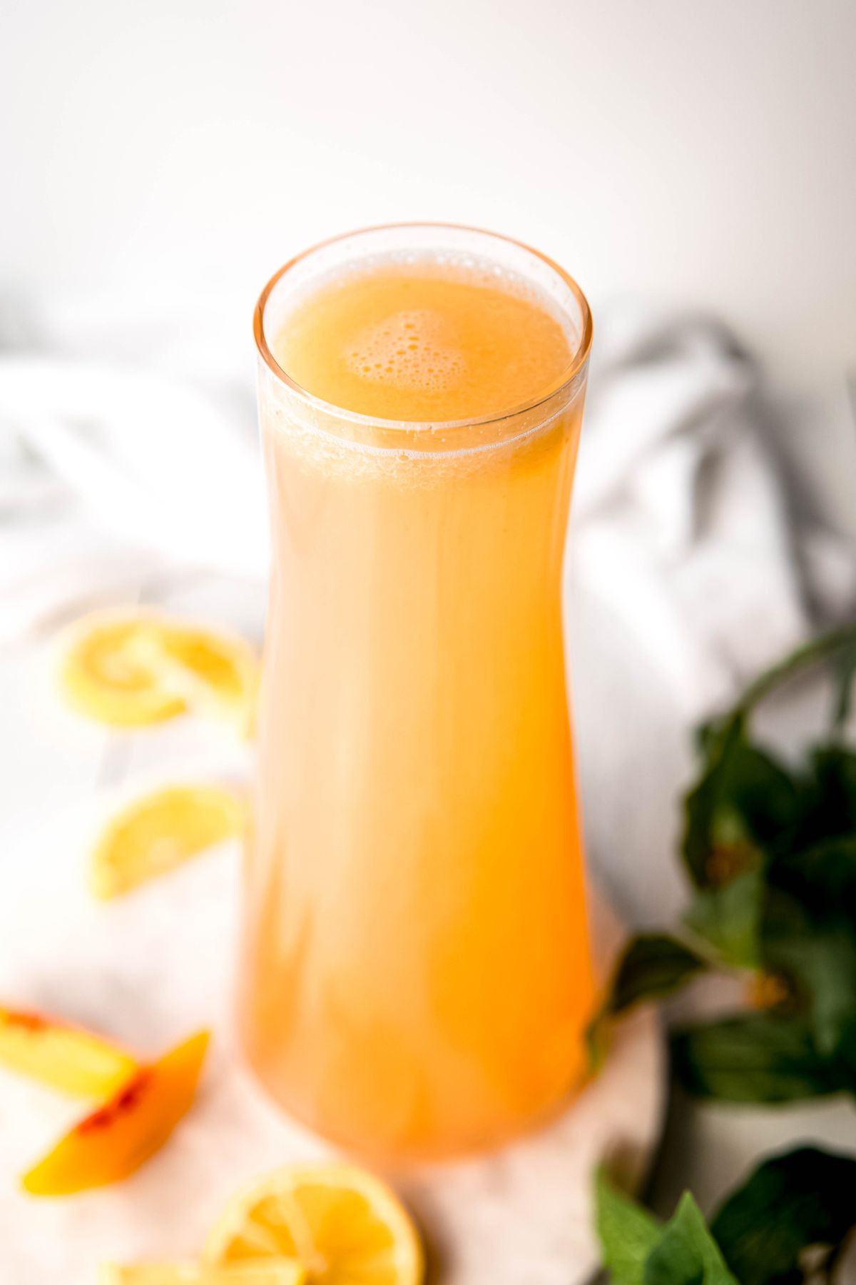 hourglass shaped glass pitcher filled to the brim with homemade peach lemonade with peach slices and lemon wheels blurred out on the white table beneath it.
