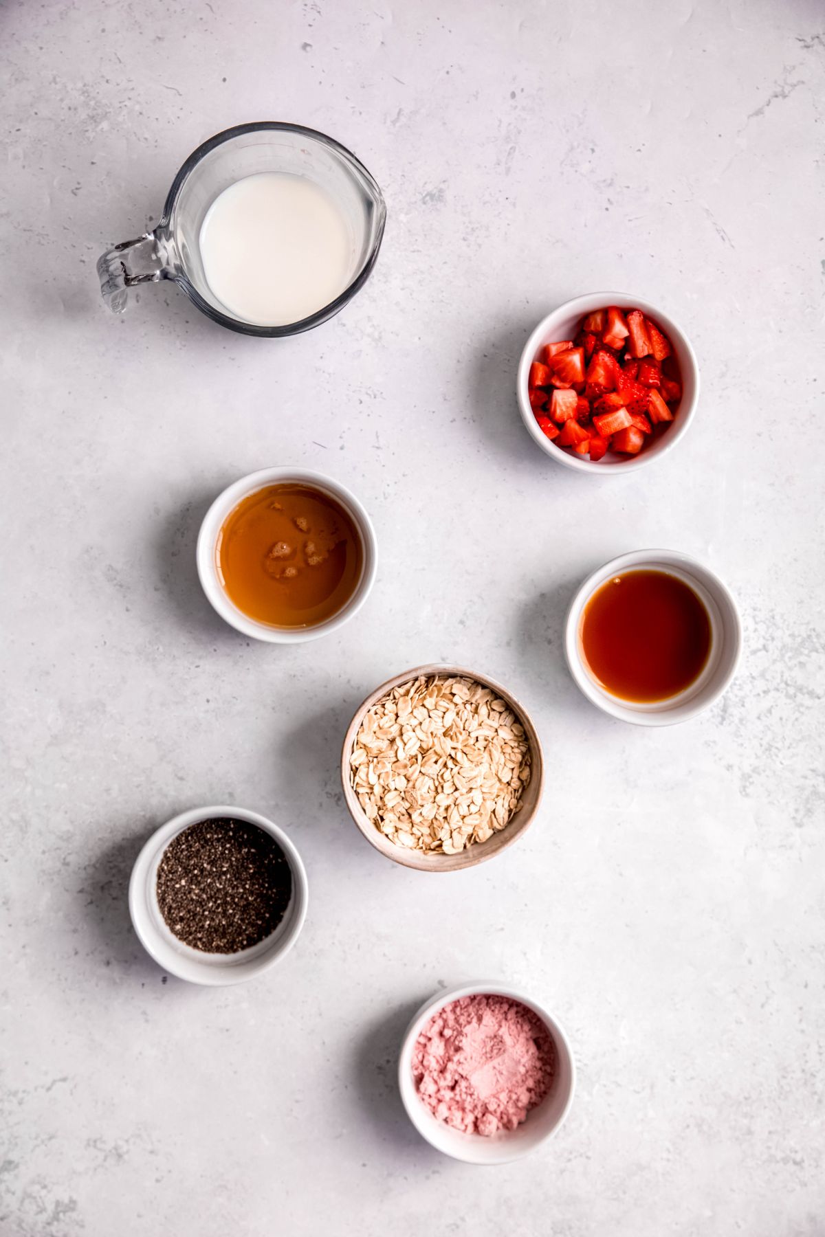 ingredients needed to make high-protein strawberry overnight oats measured out into white bowls on a white table.