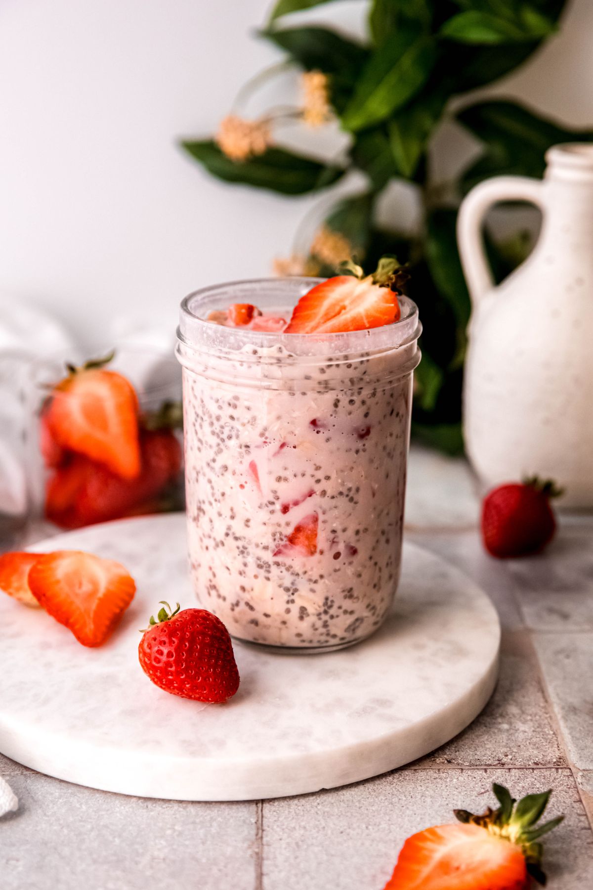jar of high protein strawberry overnight oats on a white marble round atop a grey tile table with whole and halved fresh strawberries scattered around and a ceramic white milk jug and a green houseplant blurred out behind.