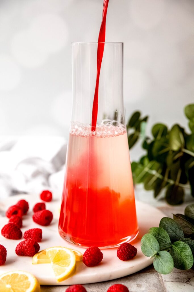 action shot of the bright red raspberry syrup being poured into the lemon water mixture, causing an ombre effect.
